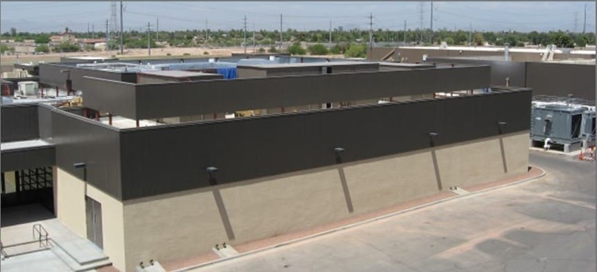 Single-story commercial or industrial building with dark upper facade, light lower walls, rooftop equipment, and a paved area in the foreground. Power lines and low landscape are visible in the background.