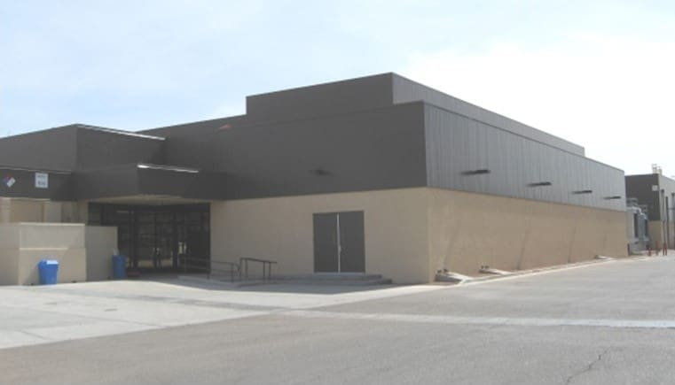 A large, modern, rectangular building with tan lower walls and dark upper walls, featuring a main entrance with glass doors and a ramp.