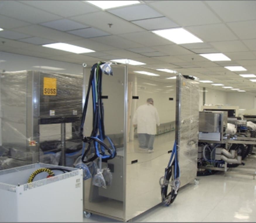 A person in a lab coat stands near large, stainless steel equipment in a brightly lit, clean laboratory or industrial facility.