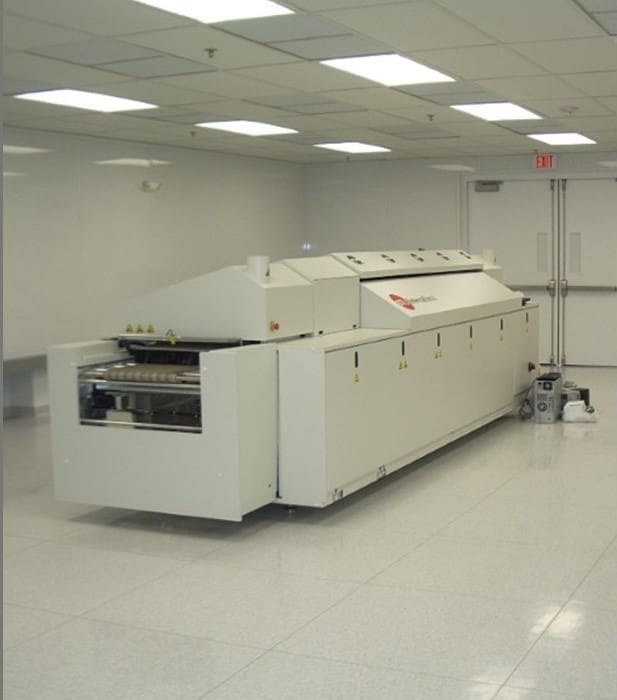 A large industrial conveyor oven sits in a clean, white room with tile floors and fluorescent lighting.