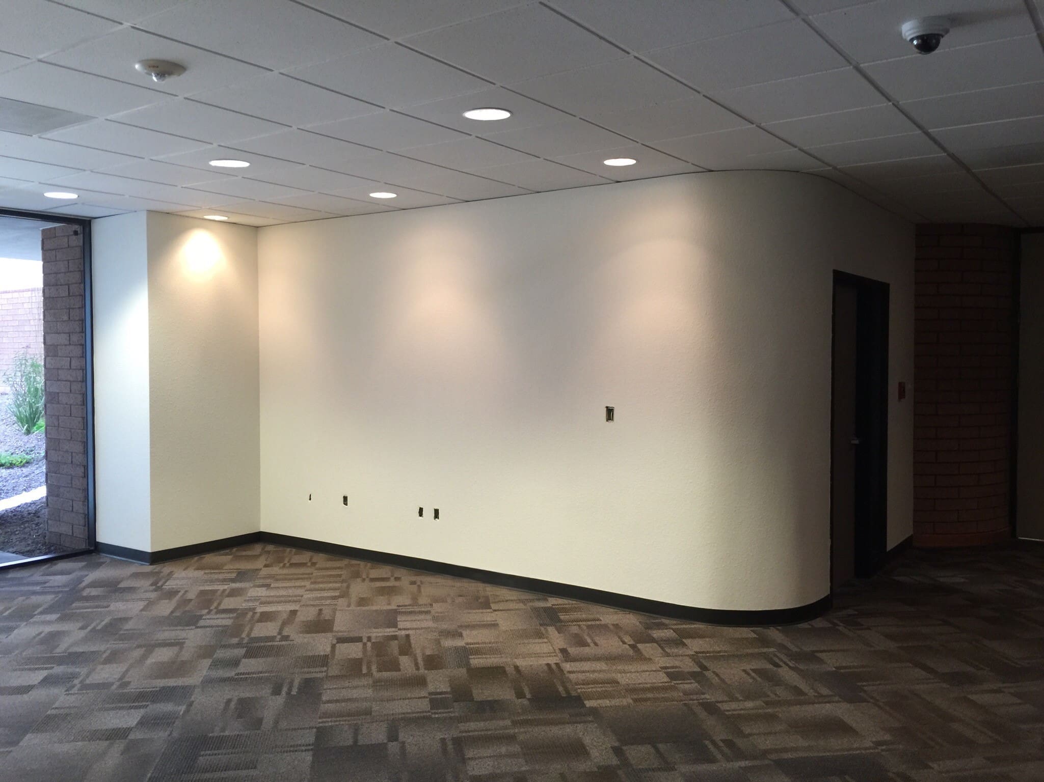 An empty office space with beige carpet, white textured walls, recessed ceiling lights, and a doorway leading to another room.