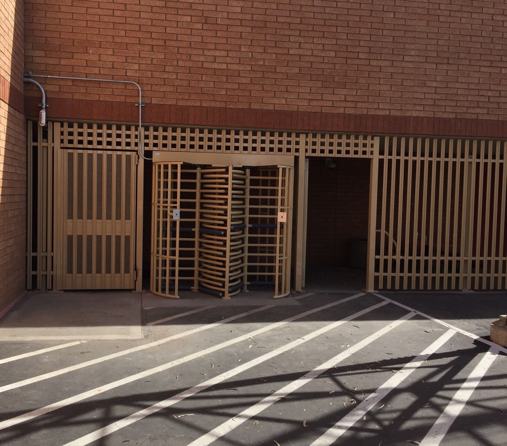 A barred security gate and rotating turnstile entrance next to a brick building, with metal fencing and striped pavement in the foreground.