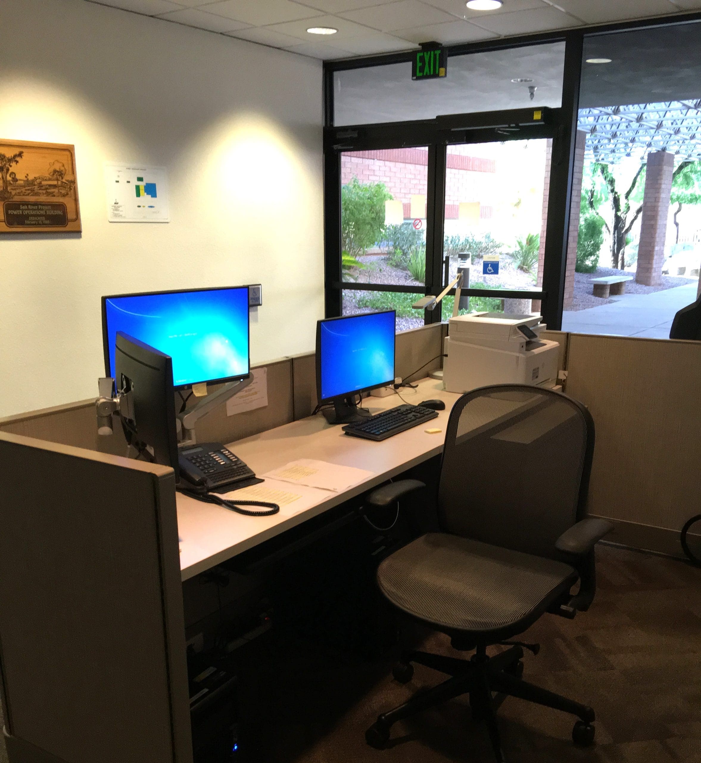 Office workstation with two monitors, a keyboard, a phone, paperwork, a printer, an empty chair, and a windowed exit door in the background.