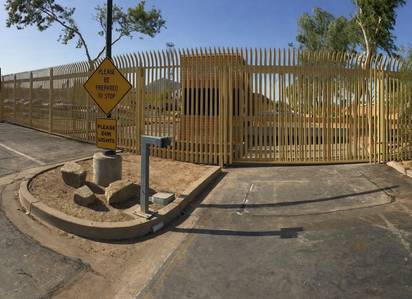 A metal security gate surrounded by a tall fence, with yellow warning signs instructing drivers to stop and dim lights; rocks and a pole are near the entrance.
