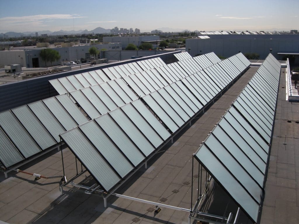 Rows of solar panels are installed on a flat rooftop in an urban area with buildings and mountains visible in the background.