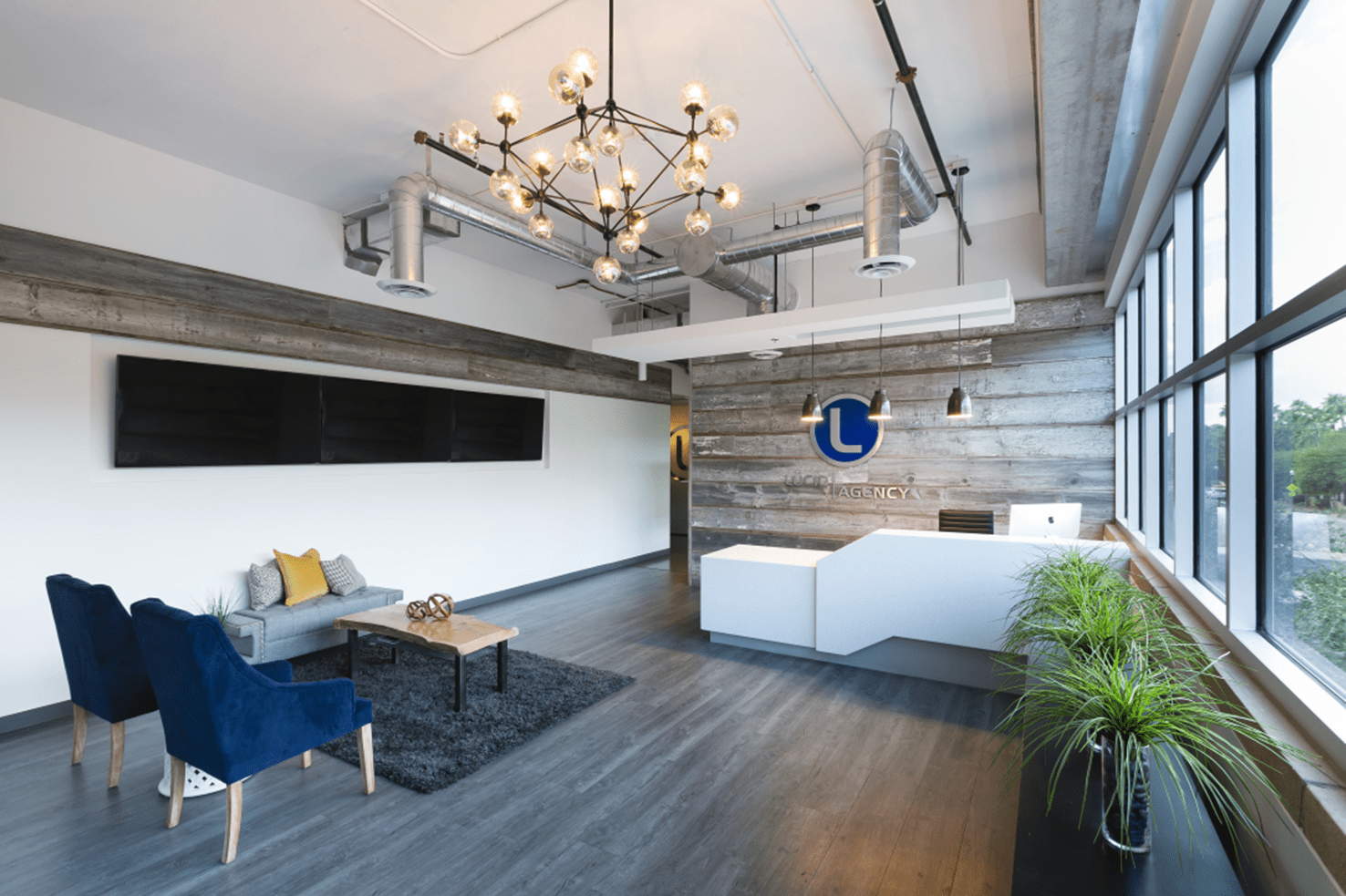 Modern office reception area with a geometric white front desk, company logo on wood-paneled wall, seating area with blue chairs, and large windows letting in natural light.