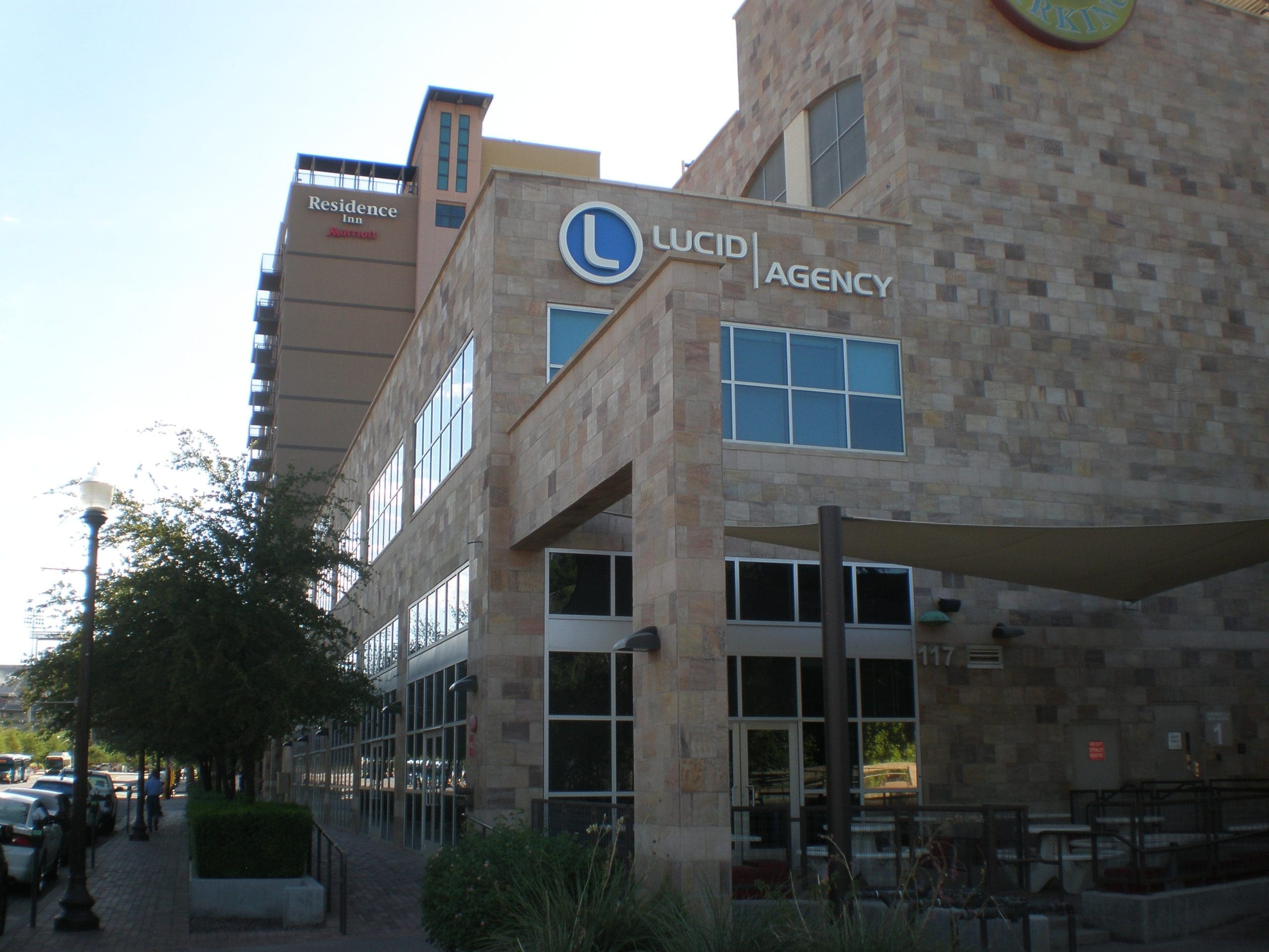 Modern, multi-story building with “Lucid Agency” signage on the stone facade; adjacent to a “Residence Inn” hotel, trees, and parked cars on the street.