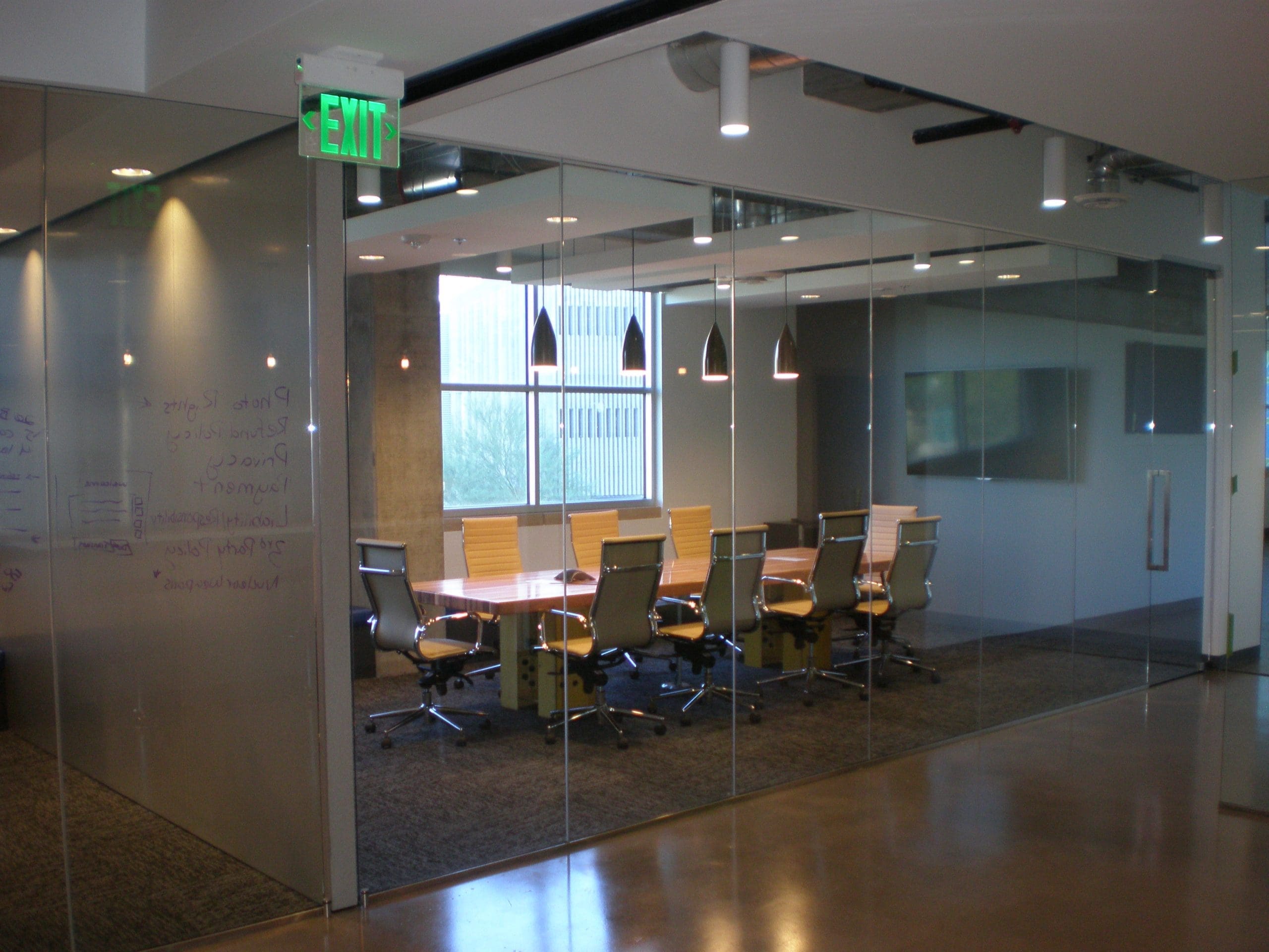 A modern glass-walled conference room with a long table, ten chairs, hanging lights, and a large monitor on the wall. An exit sign is visible near the entrance.