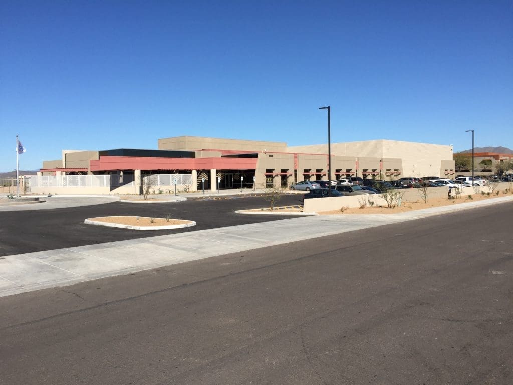 A large, modern one-story building with a parking lot in front, surrounded by desert landscaping under a clear blue sky.