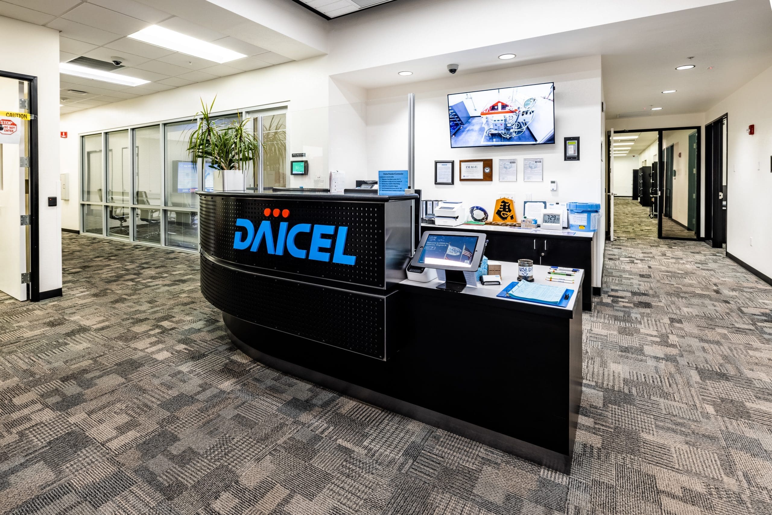 Reception area of an office with a curved desk displaying the DAICEL logo, office equipment, wall-mounted TV, framed certificates, and glass-walled meeting rooms in the background.