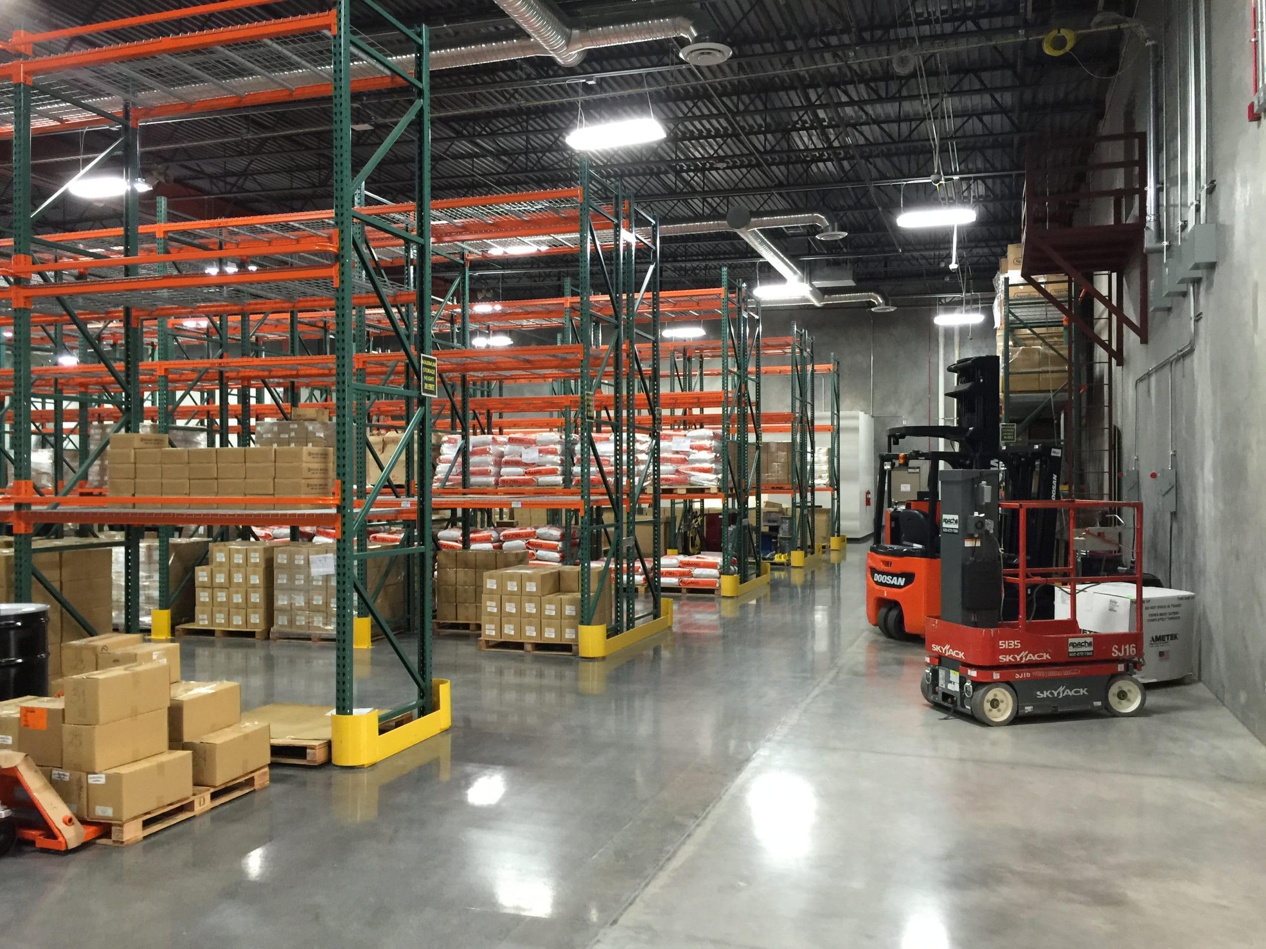 Warehouse interior with empty and partially filled shelves, stacked boxes on pallets, forklifts, and polished concrete floor.
