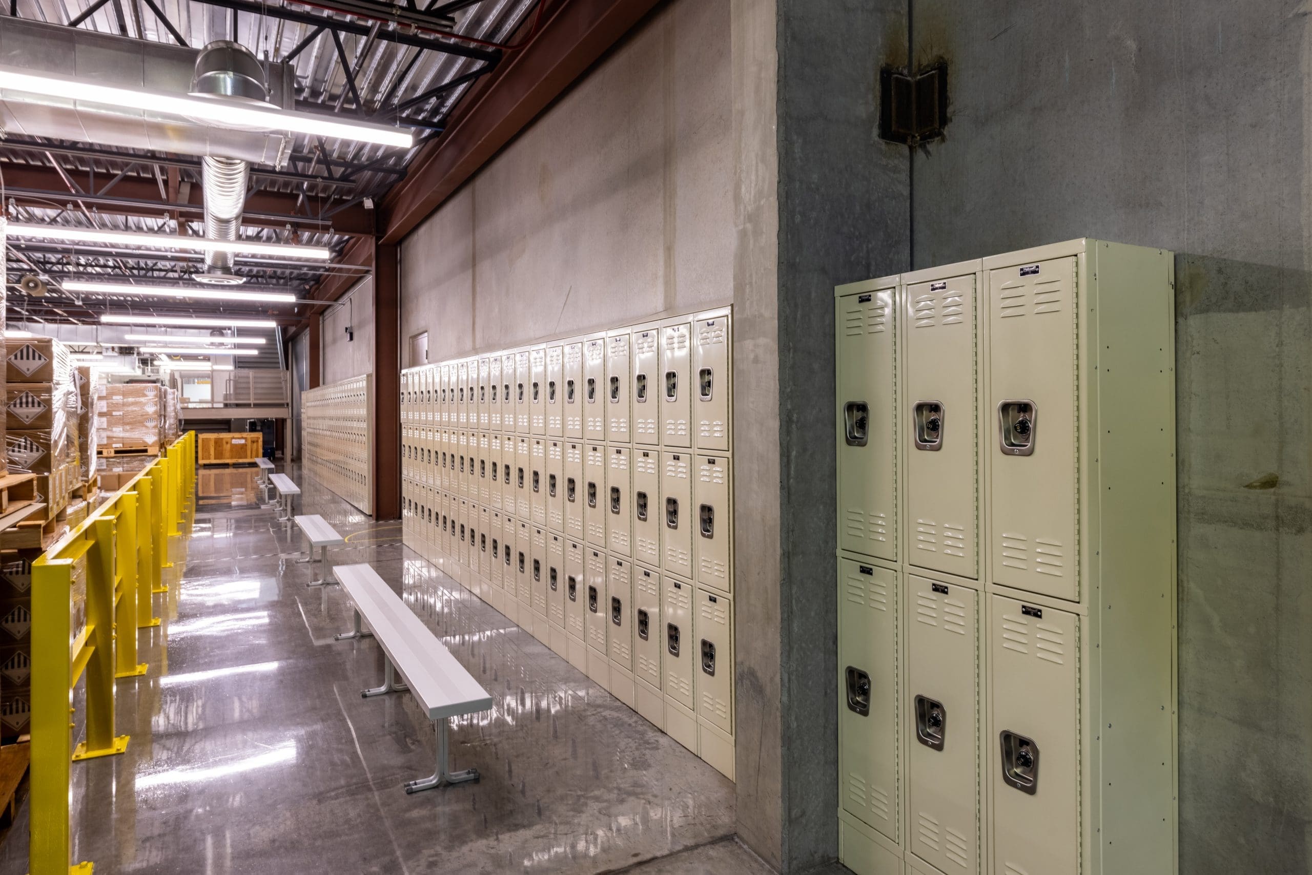 Rows of metal lockers line a concrete wall in a brightly lit industrial locker room with benches and high ceilings.