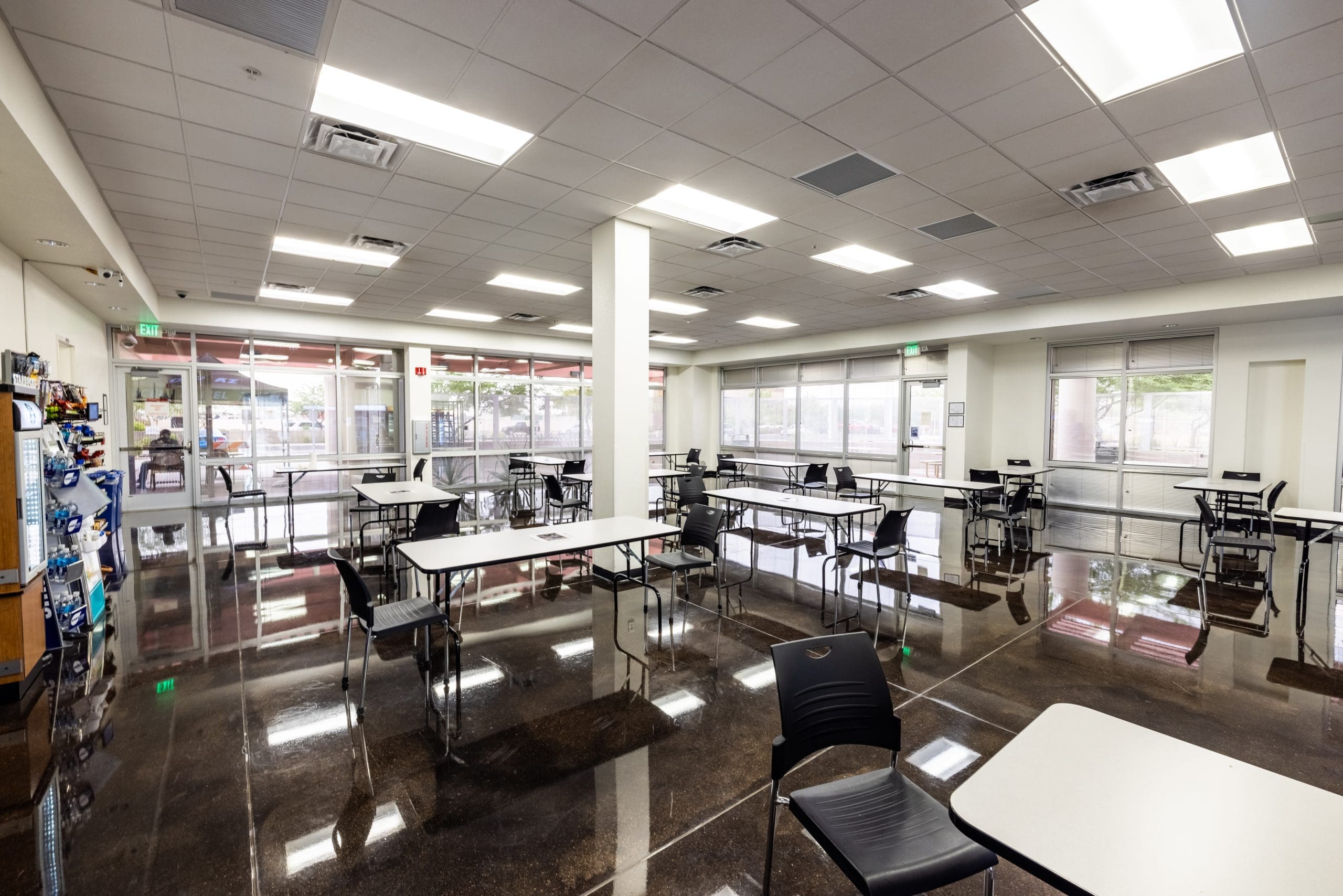 A clean, empty cafeteria with black chairs, white tables, vending machines on the left, large windows, and overhead fluorescent lighting.