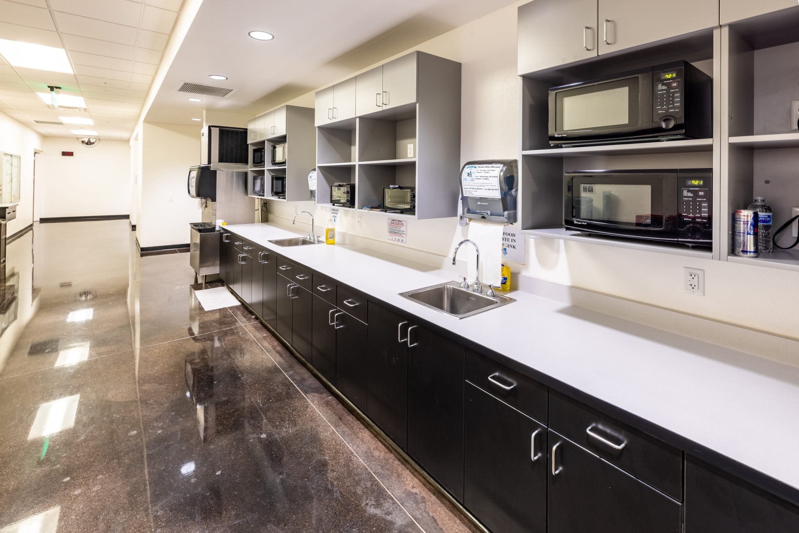 A break room kitchenette with multiple microwaves, a sink, open shelves, cabinets, soap dispensers, and a polished floor.