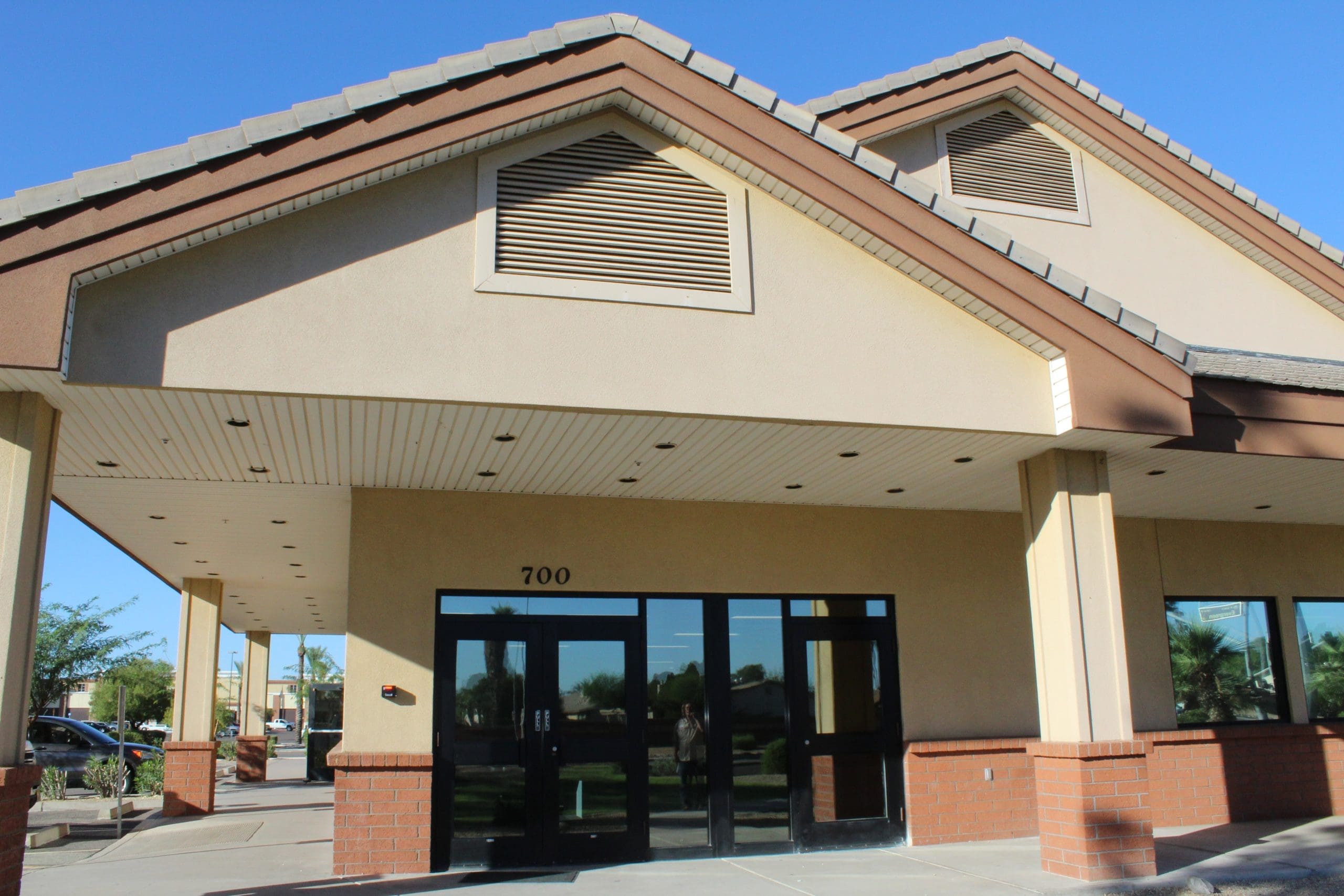 Single-story beige and brown building with a covered entrance and glass double doors, marked with the number 700 above the door, photographed on a clear sunny day.