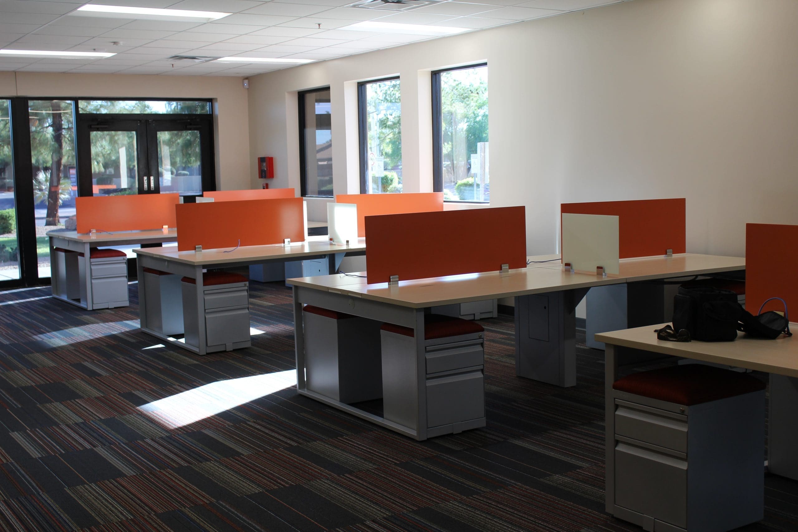 Modern office space with multiple desks arranged in rows, each with orange dividers, on a carpeted floor near large windows.