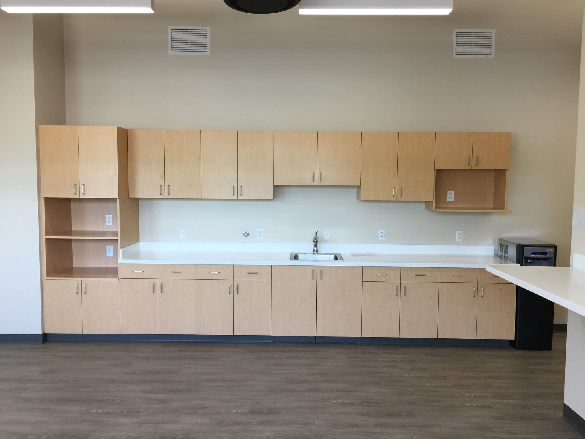 A kitchenette with light wood cabinets, a white countertop, a sink, a microwave, and a small safe on the right, set against a beige wall with a wood-style floor.