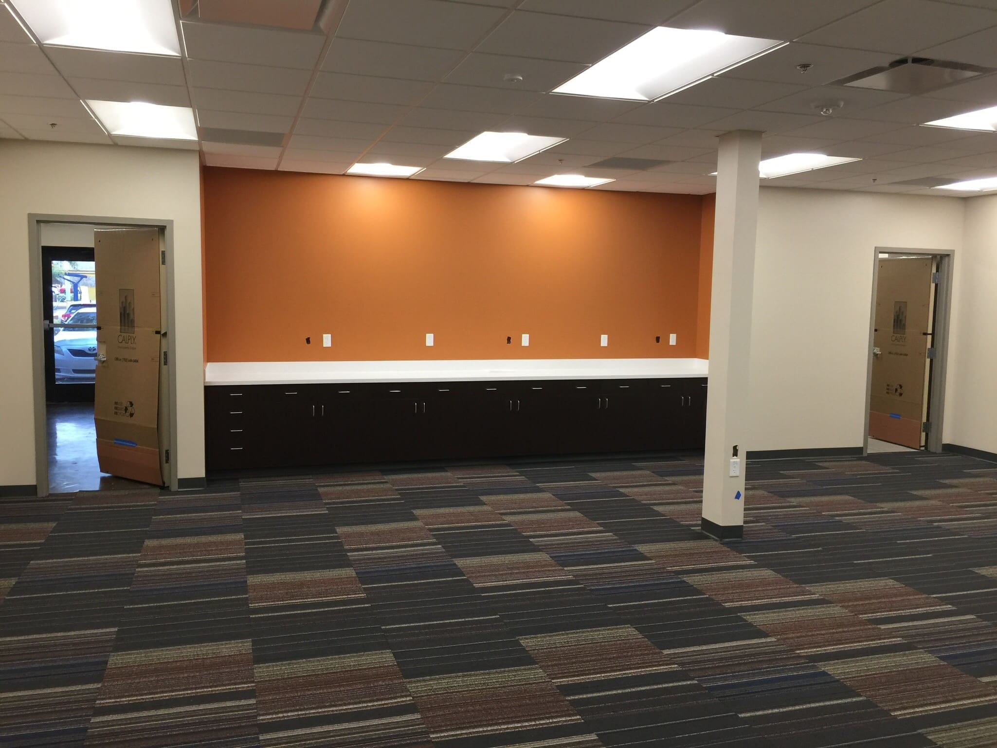 Office space with patterned carpet, an orange accent wall, dark cabinets with a white countertop, and two doors on either side. Ceiling lights are on.