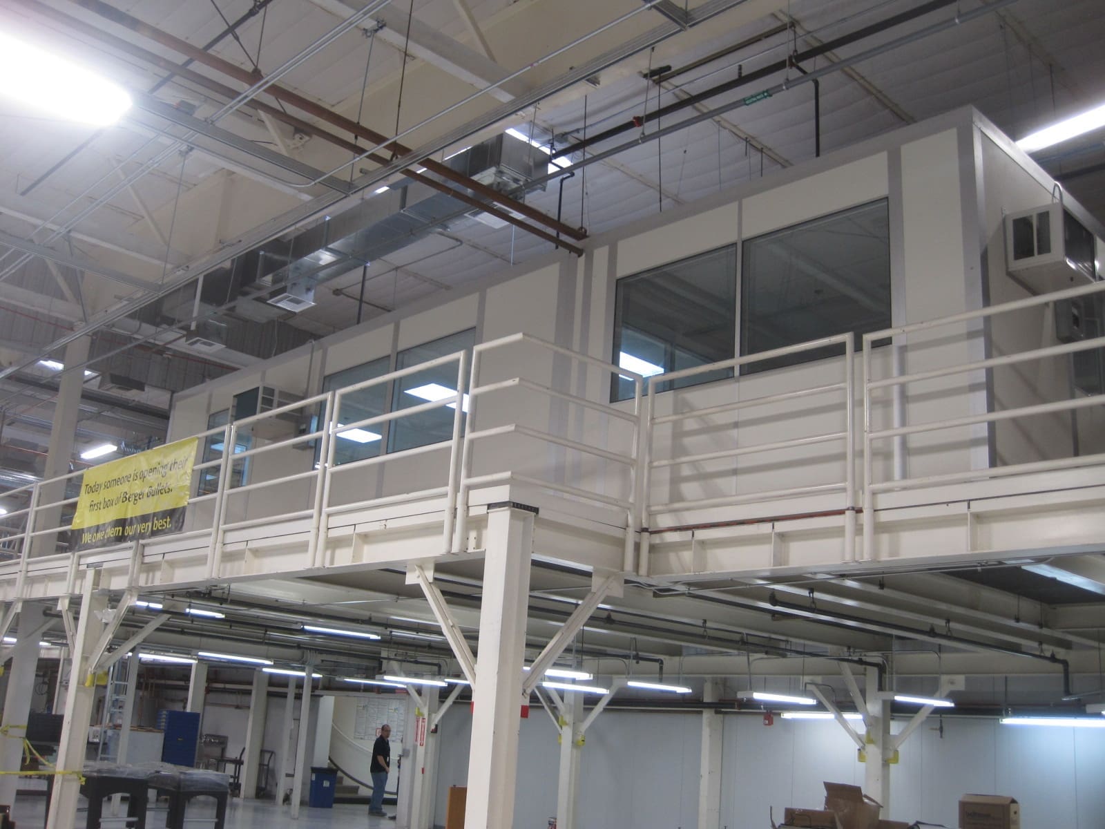 Elevated enclosed office space with large windows and railing inside an industrial warehouse setting.