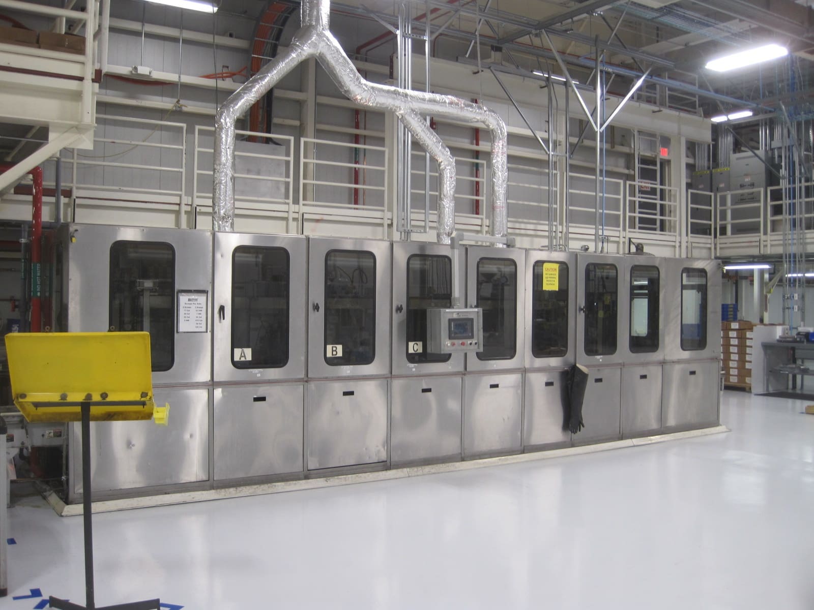 Long stainless steel industrial machine with multiple closed compartments and overhead ventilation ducts in a clean, modern manufacturing facility.