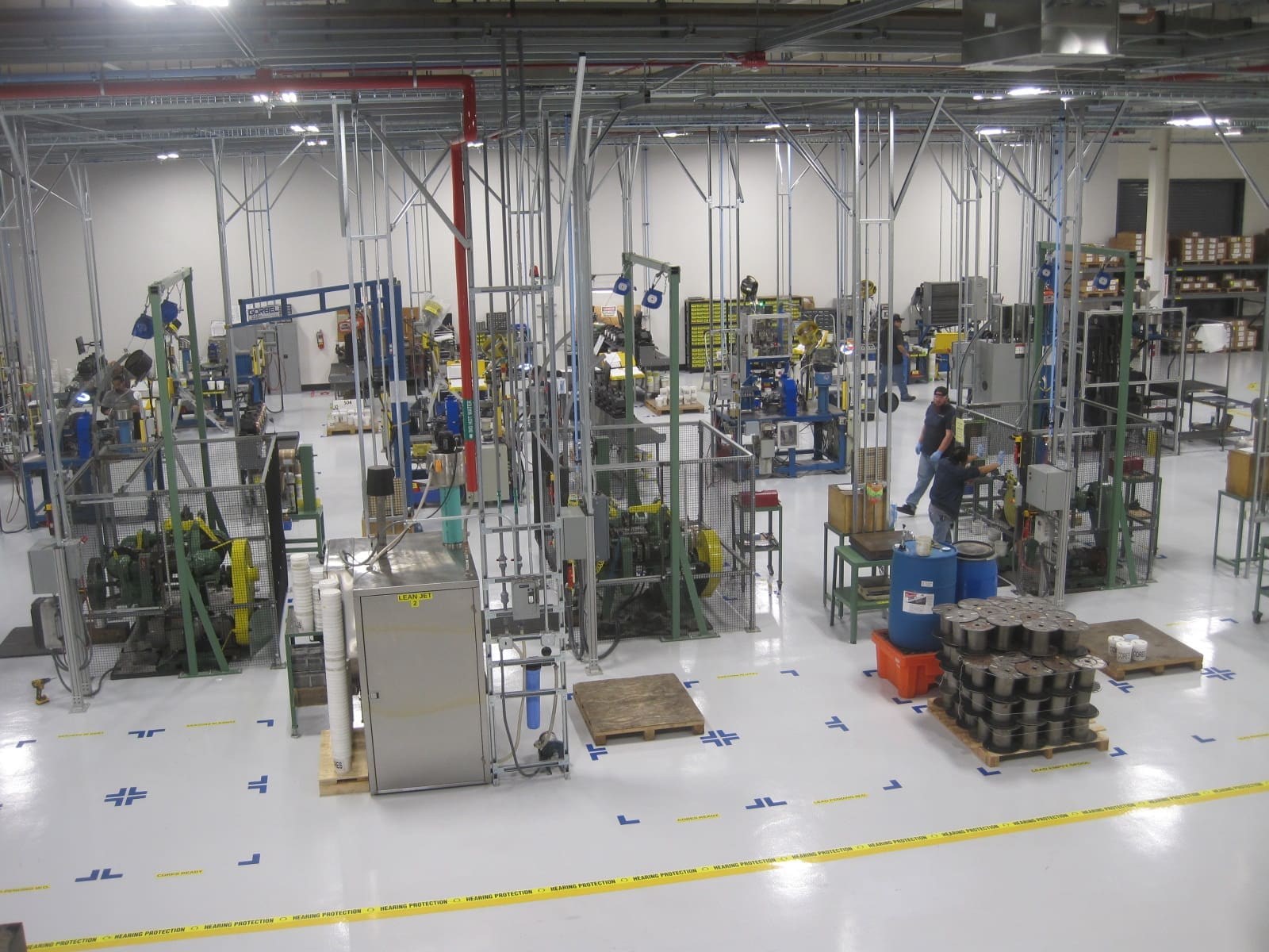 Large industrial factory floor with various machines, metal structures, and workers operating equipment, organized in designated areas with safety markings on the floor.