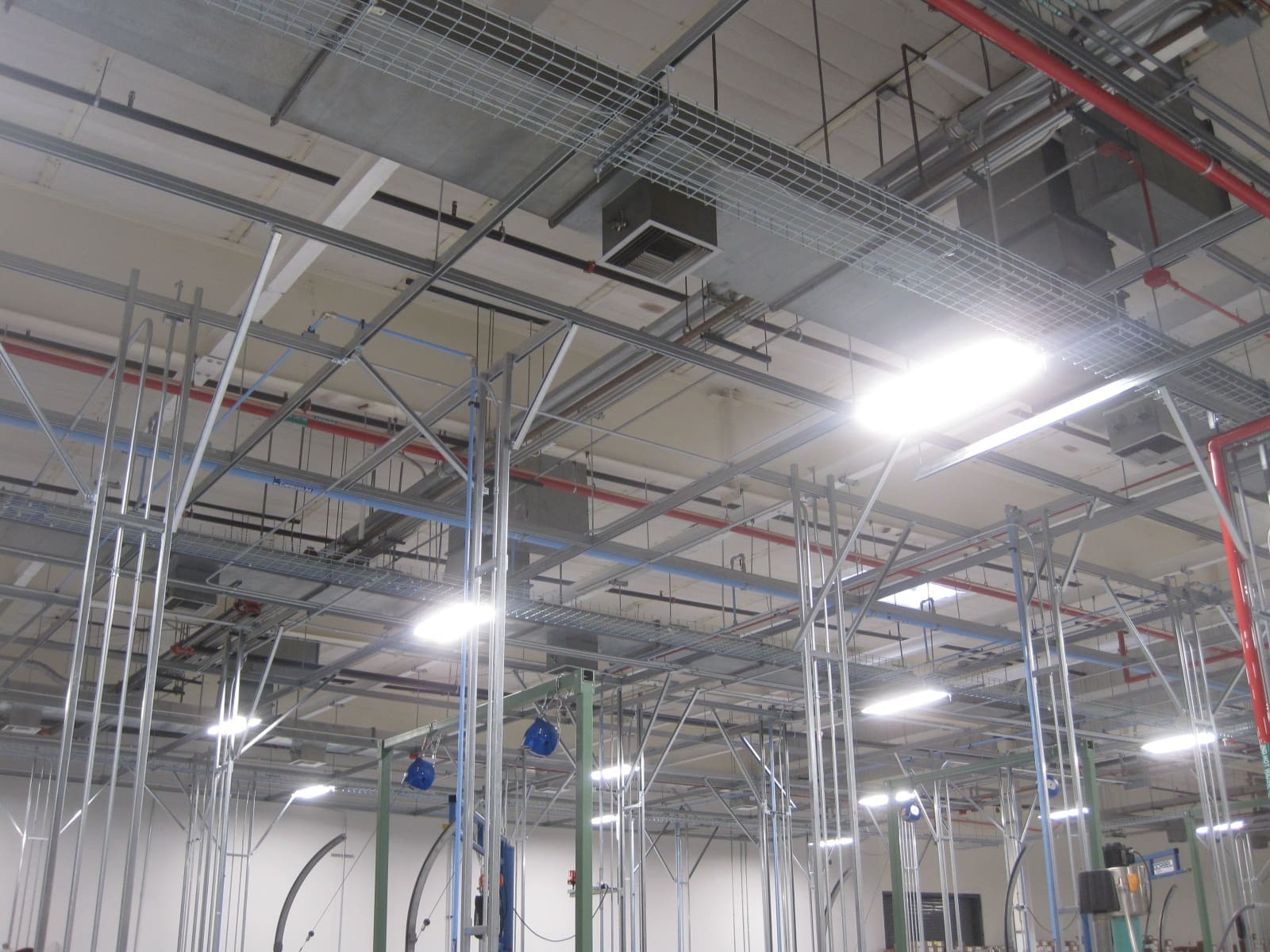 Industrial ceiling with exposed ductwork, metal framework, electrical conduits, and hanging fluorescent lights in a large indoor facility.
