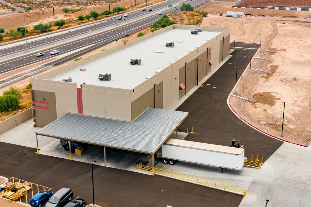 Aerial view of a large, beige industrial warehouse with a loading dock and a semi-trailer parked under a metal awning. The building is surrounded by asphalt and desert landscape.