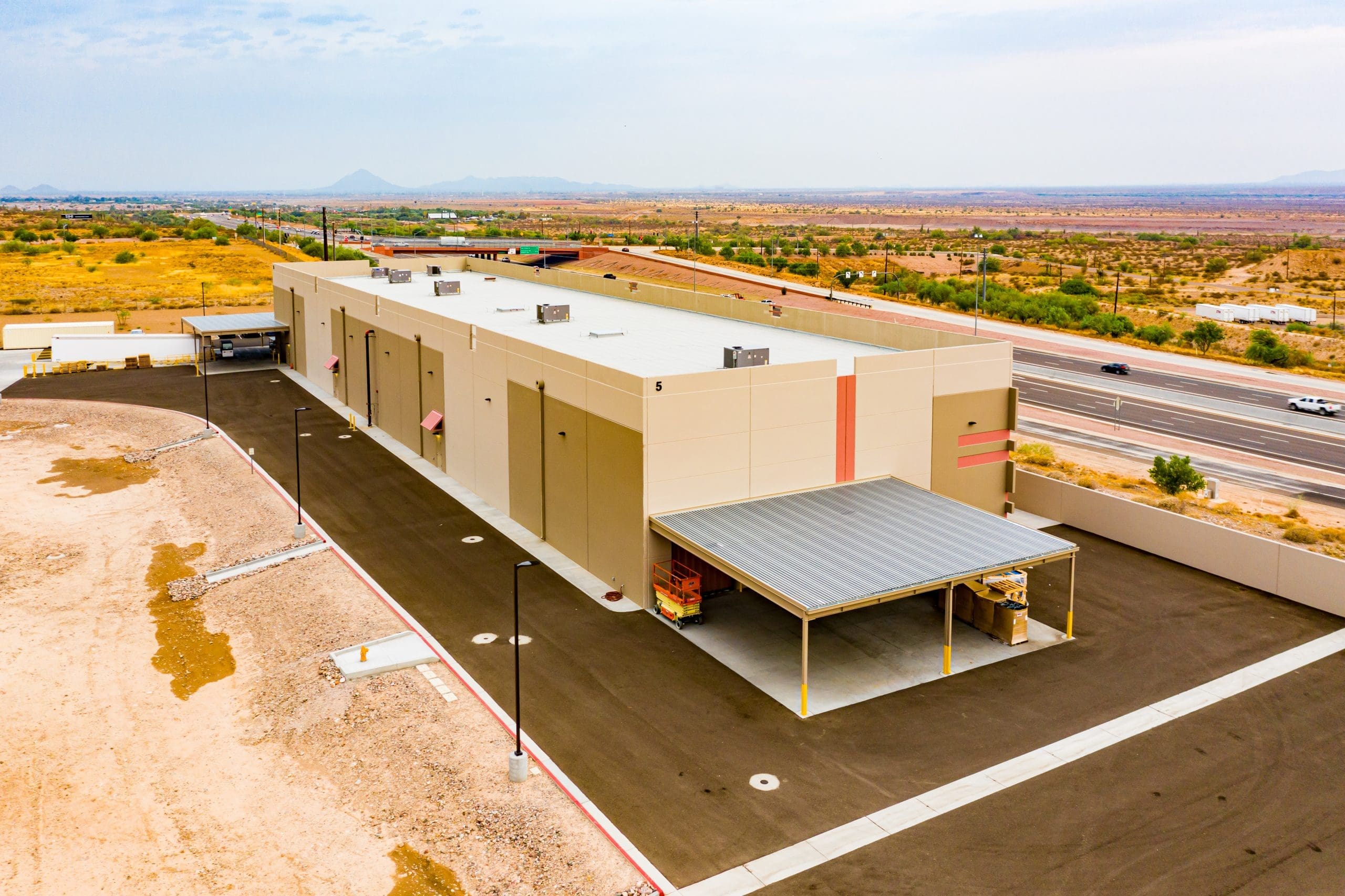 Aerial view of a large beige industrial warehouse with a covered loading area, surrounded by paved roads and desert landscape.