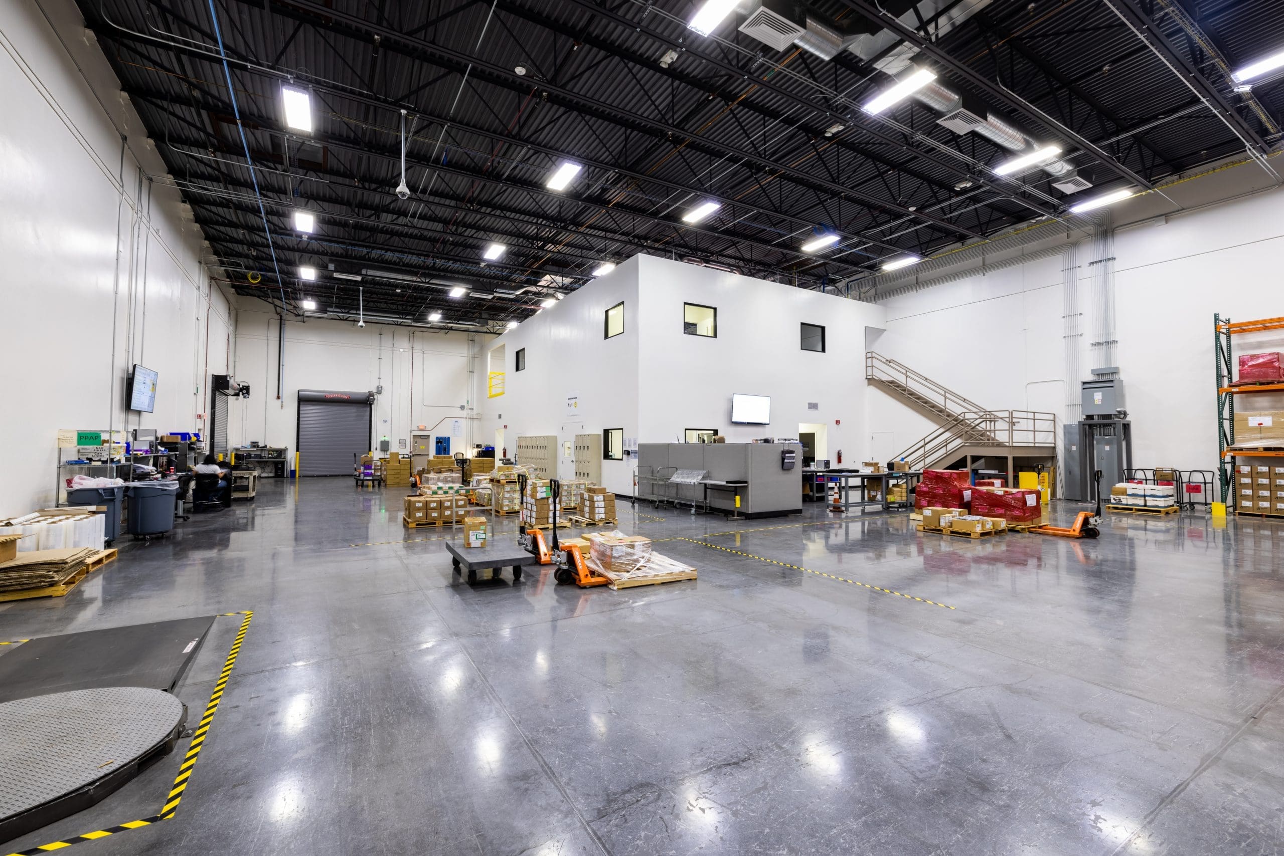 Large warehouse interior with polished concrete floor, stacks of pallets, carts, and shelving; enclosed office area with windows elevated at the back.