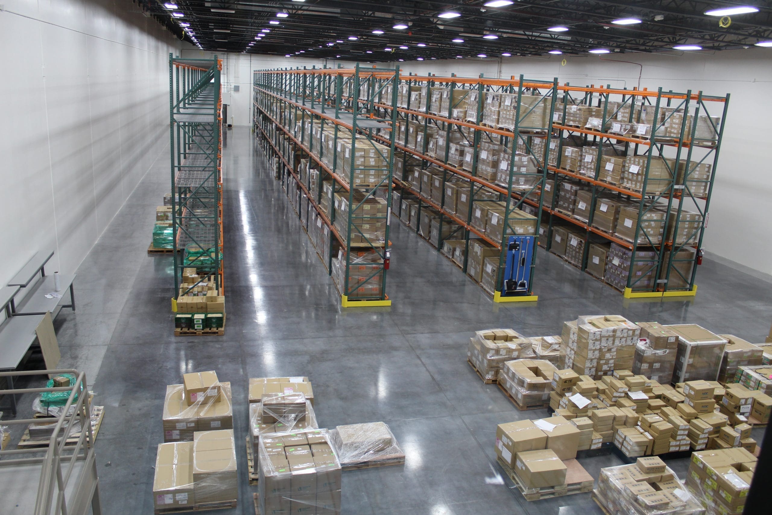 Large warehouse interior with tall shelving racks filled with boxes and pallets; additional boxes and pallets are stacked on the polished concrete floor.