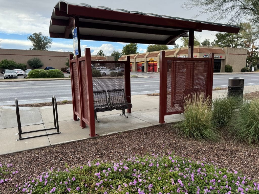 A covered bus stop shelter with two benches, a bike rack, and a trash can, located near a road and shopping center with storefronts in the background.