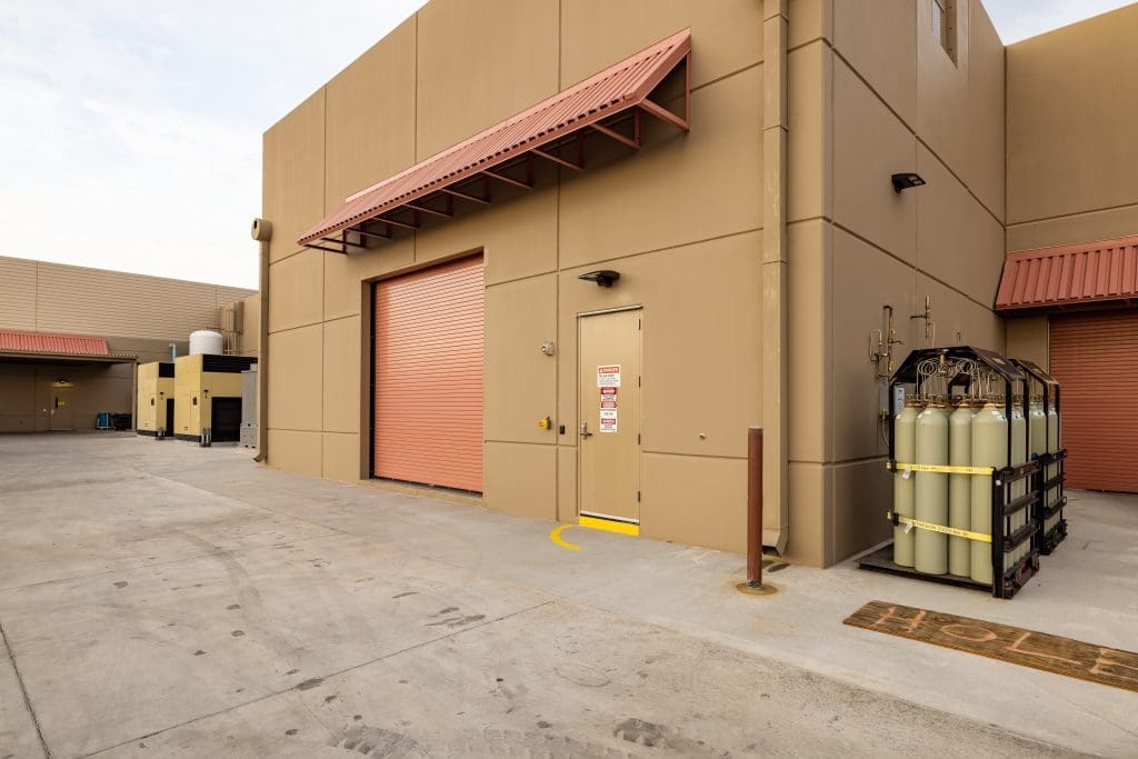 Industrial building exterior with tan walls, a closed red roll-up door, a smaller door with warning signs, and gas cylinders stored beside the building on a concrete surface.