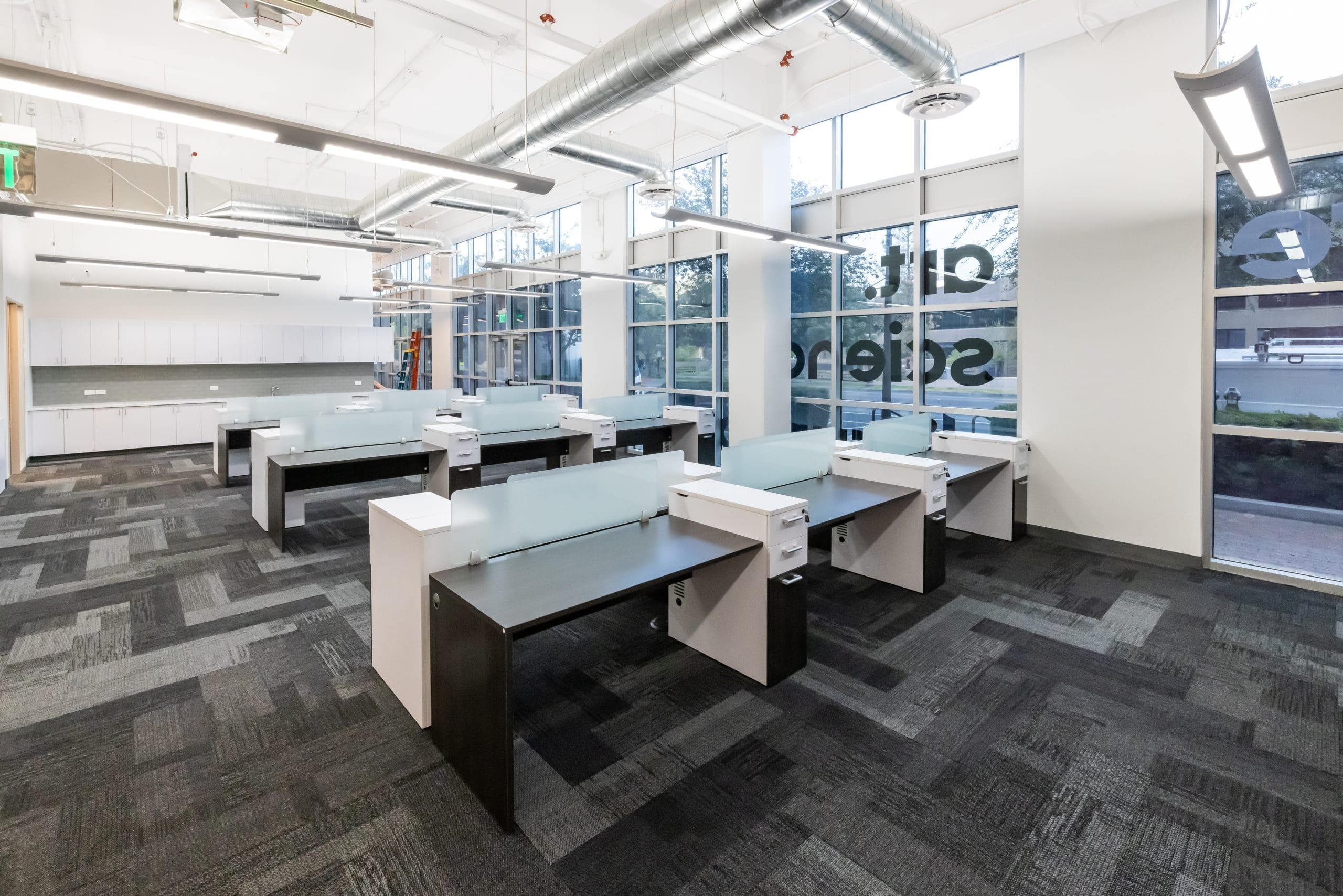 Modern open office space with multiple empty workstations, large windows, overhead lighting, and exposed ductwork on the ceiling.
