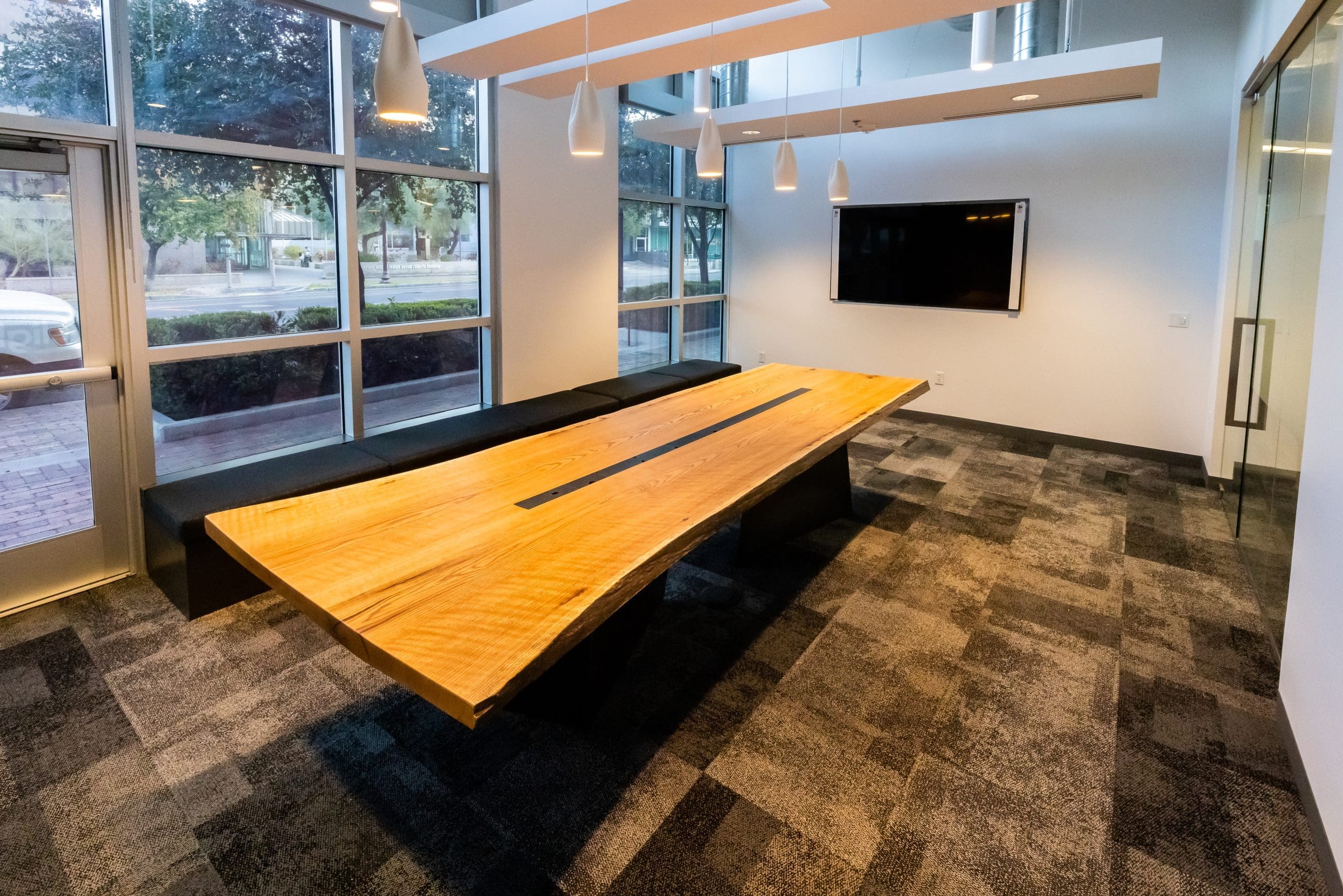 A modern conference room with a large wooden table, black benches, a wall-mounted TV, pendant lights, and floor-to-ceiling windows.