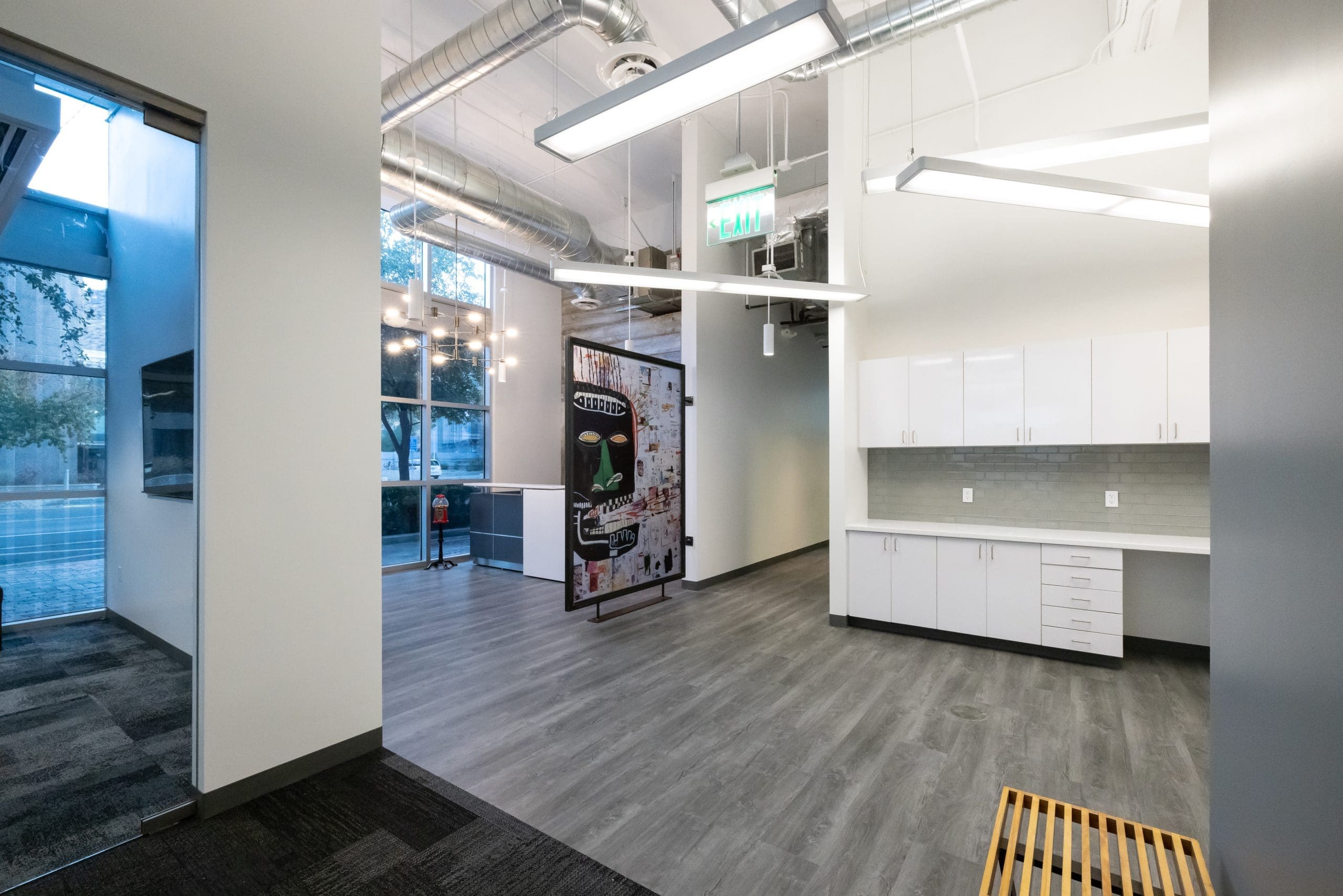 A modern office interior with gray flooring, white cabinets, exposed ductwork, large windows, and a glass door leading outside.