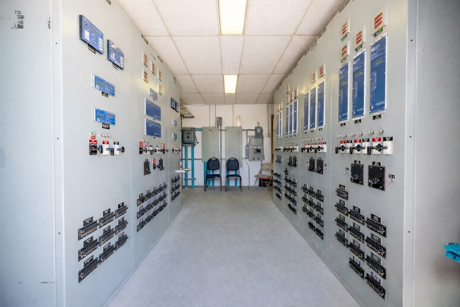 A control room with two rows of electrical panels, switches, and monitors lining the walls, leading to two chairs and more equipment at the far end.