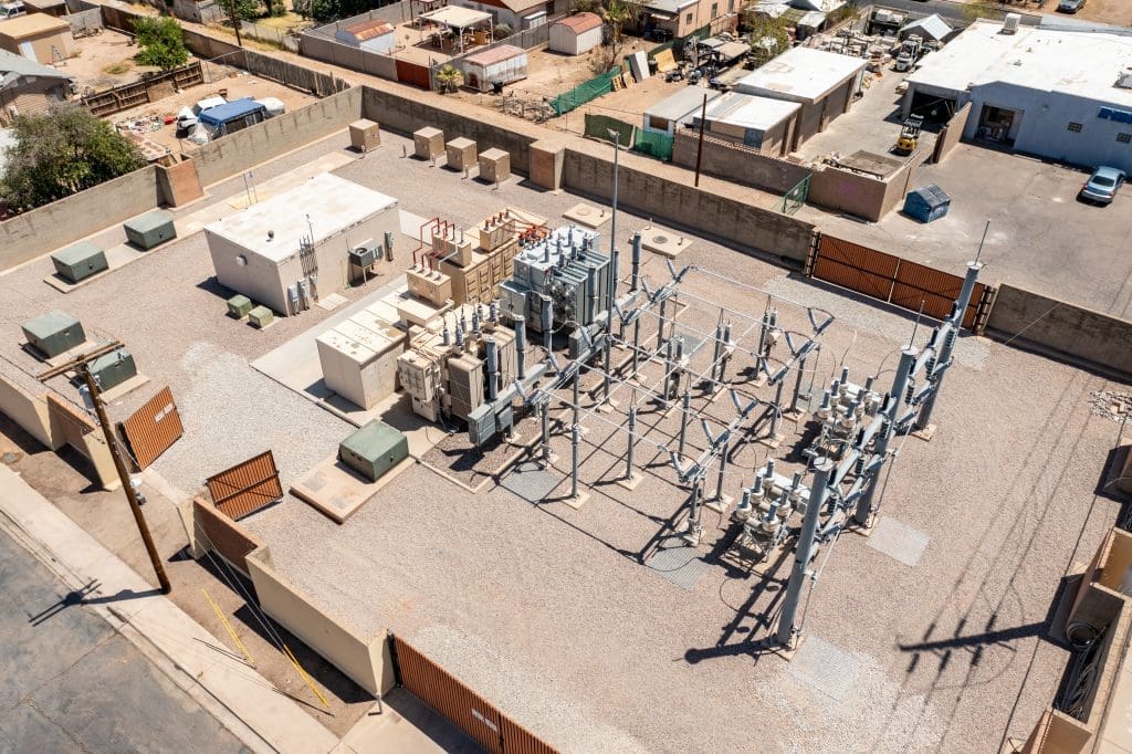 Aerial view of an electrical substation with transformers, metal structures, and equipment surrounded by a wall in an urban area.