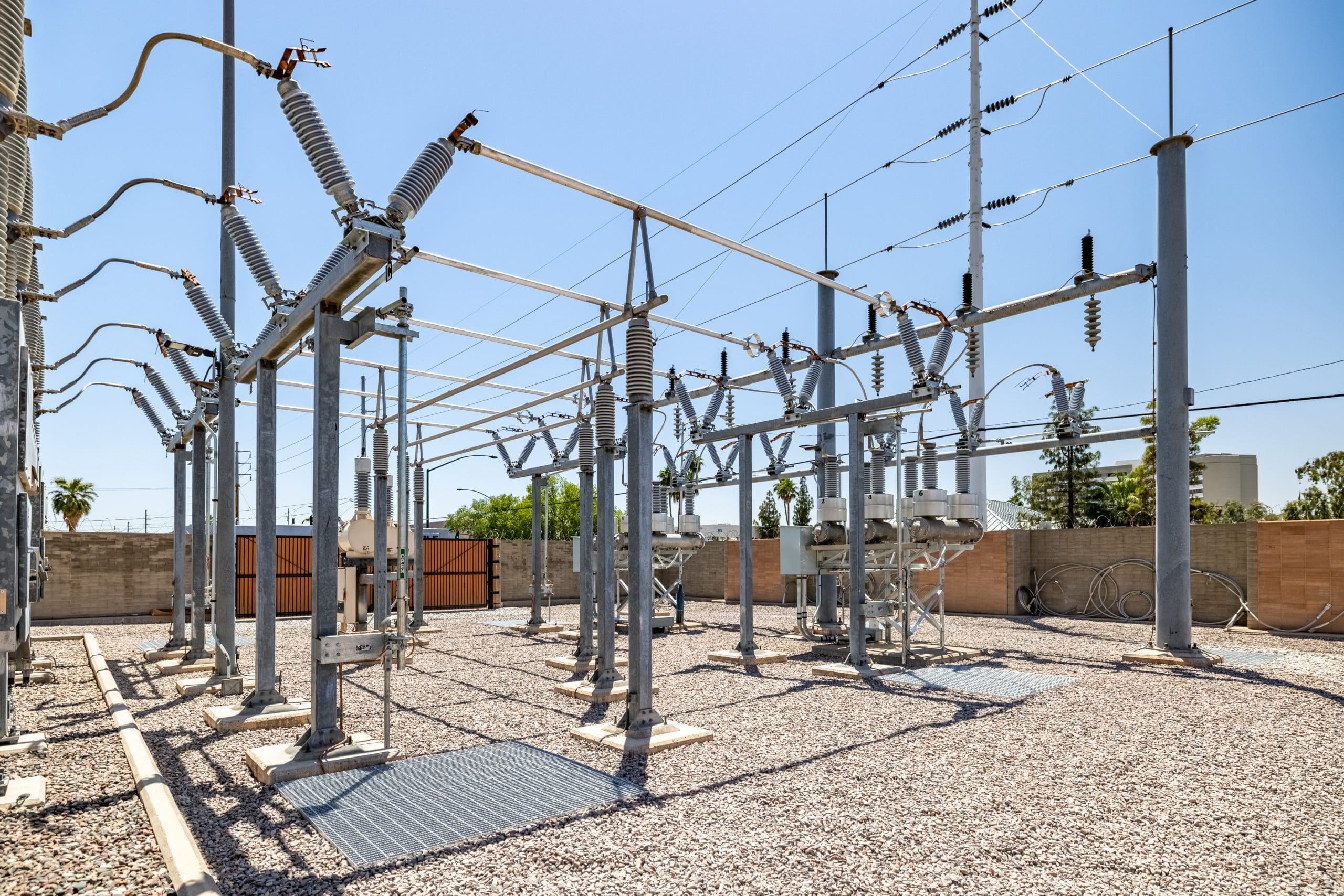 Outdoor electrical substation with metal structures, insulators, and power lines on a gravel surface, set against a clear sky and fenced perimeter.