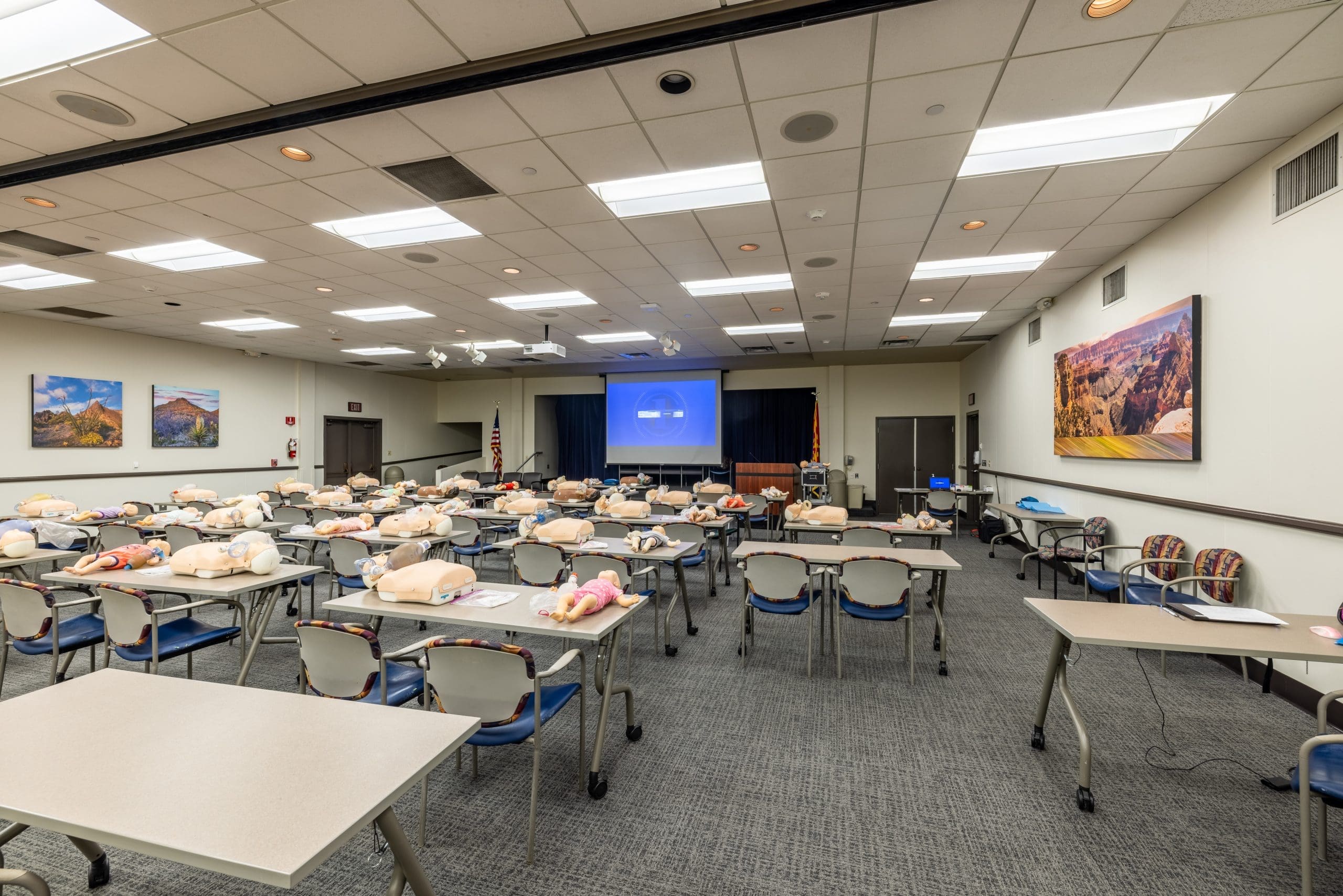 A classroom setup with rows of tables and chairs, each with a CPR training mannequin; a projector screen at the front displays a blue image.