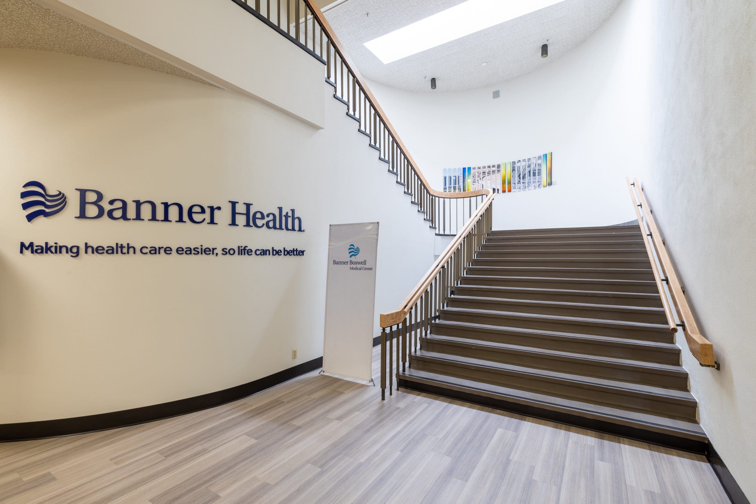 Staircase inside a building with a Banner Health sign and slogan on the wall, a standing Banner Health sign, and artwork visible at the top of the stairs.