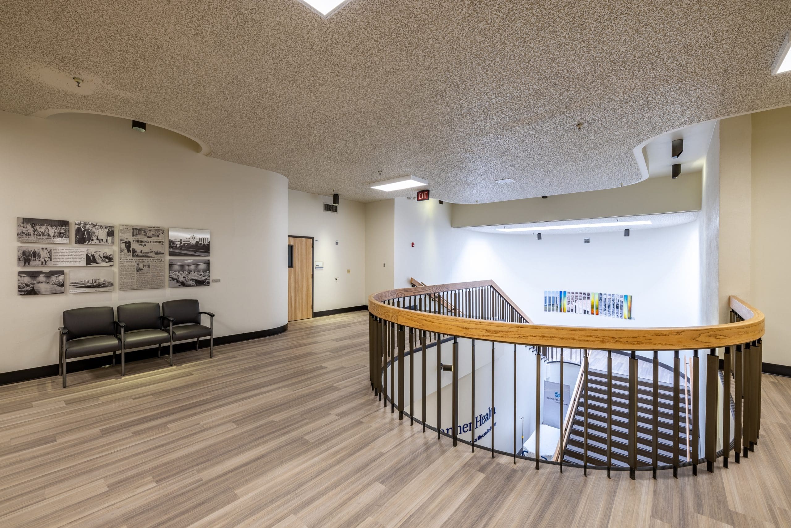 Second-floor hallway with wood railings around a staircase, seating area with three chairs, wall-mounted photos, and light wood flooring.