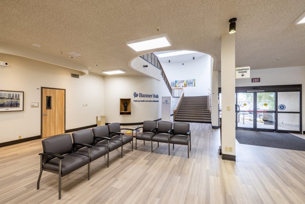 A medical office waiting area with rows of black chairs, a staircase, a sign reading "Banner Health," and natural light from windows by the entrance.