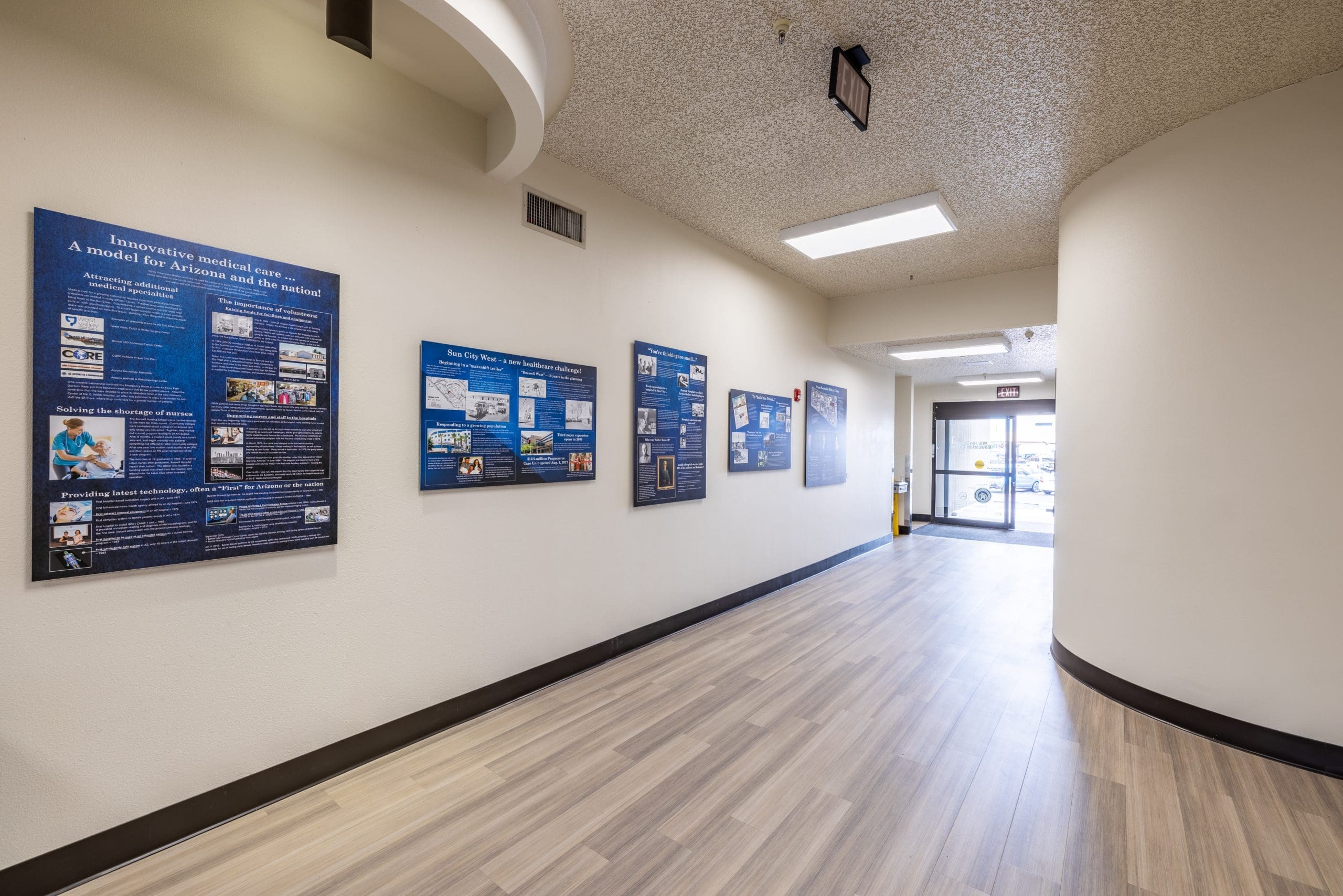 A hallway with wood-look flooring and display boards featuring informational posters on the left wall, leading to a glass exit door.
