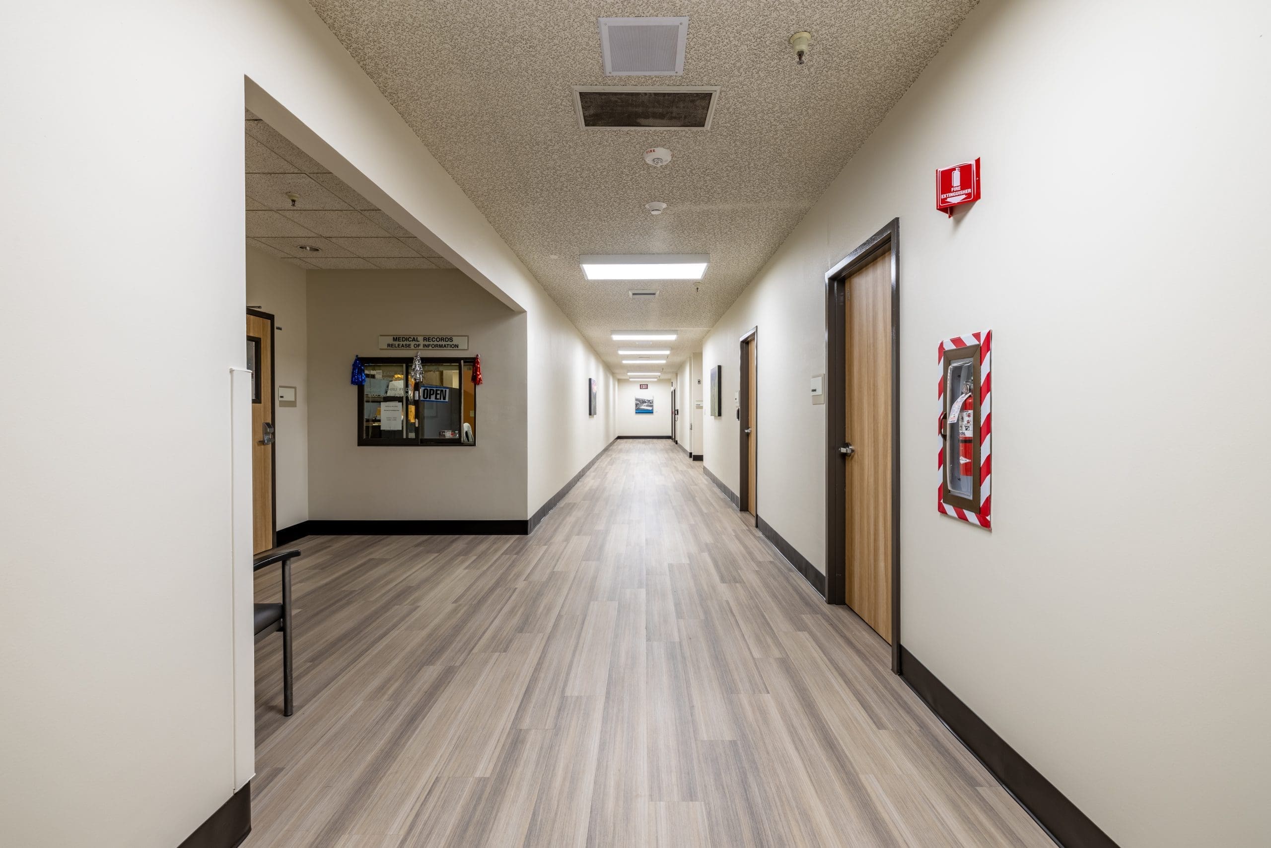 Long, empty hallway in an office or school building with closed wooden doors, beige walls, light wood flooring, a fire extinguisher, and a bulletin board.