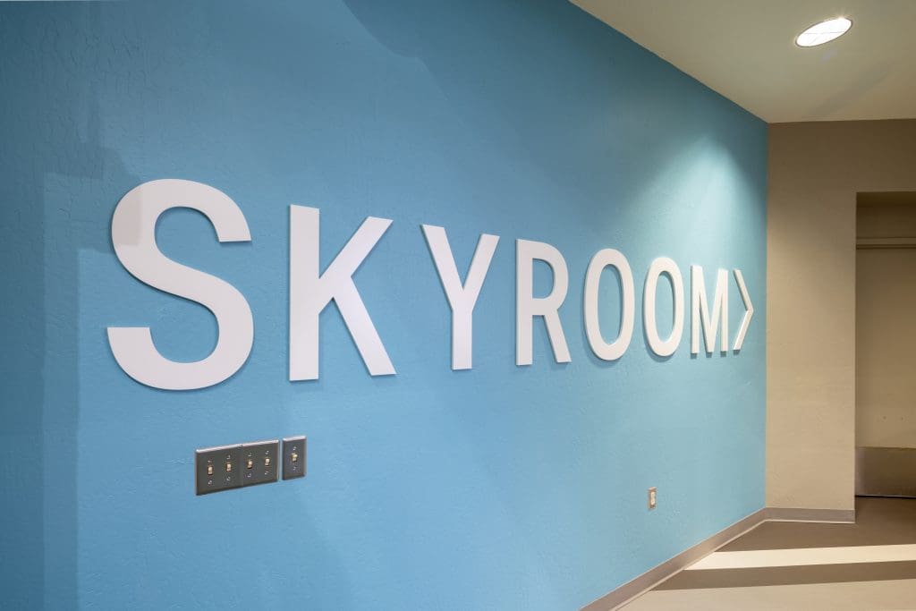 Large white letters spelling "SKYROOM" are mounted on a light blue wall with an arrow pointing right.