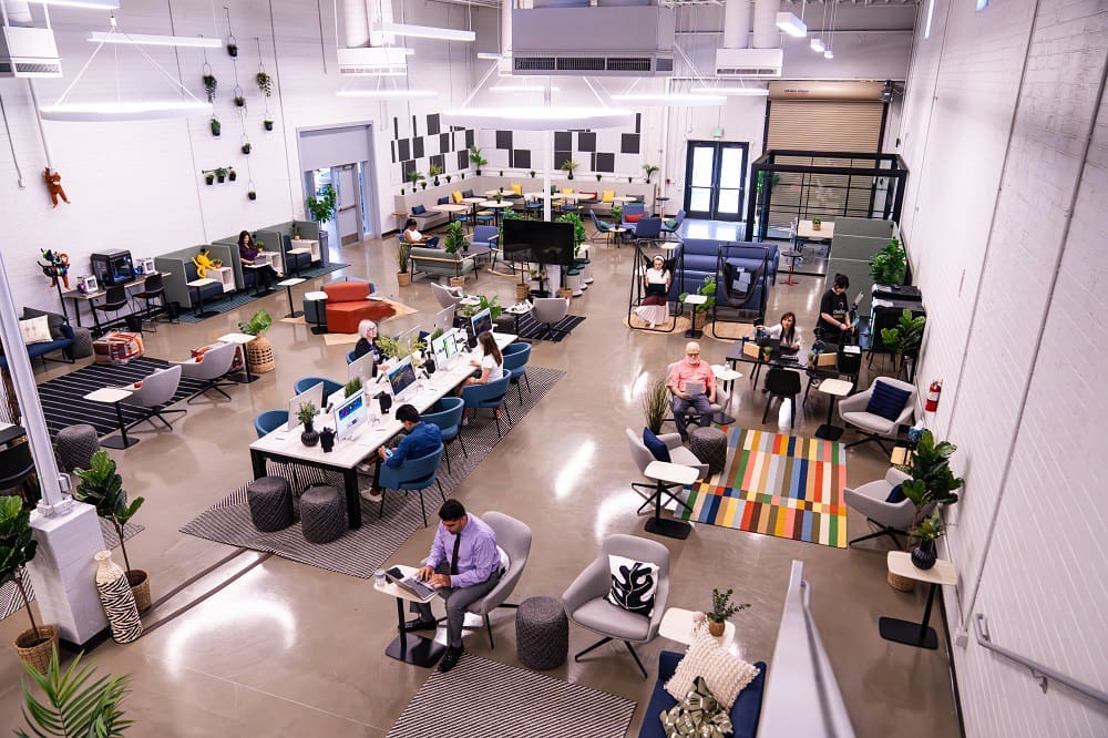 Spacious, modern coworking office with people working at desks, sofas, and tables; bright lighting, plants, and colorful decor throughout the open space.