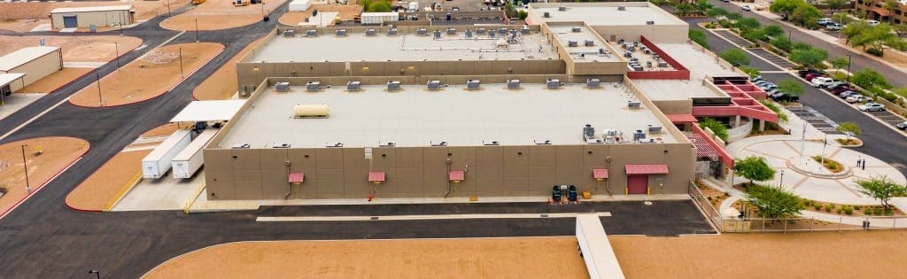 Aerial view of a large industrial building with a flat roof, loading docks, surrounding parking lots, and landscaped areas in a desert environment.