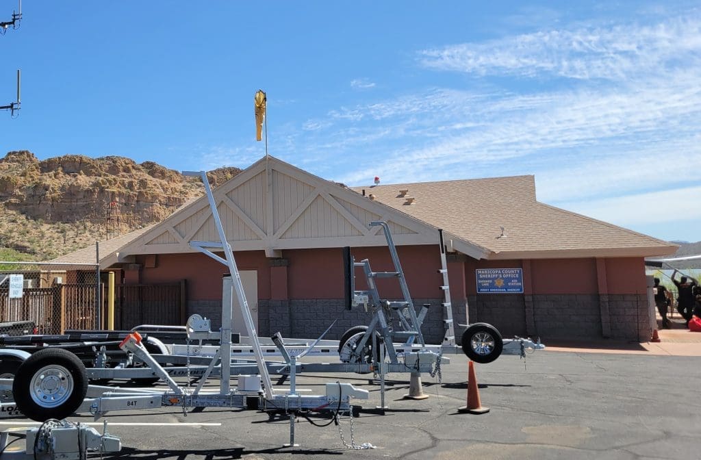 A single-story building with a peaked roof, several boat trailers parked in front, and rocky hills in the background under a blue sky.