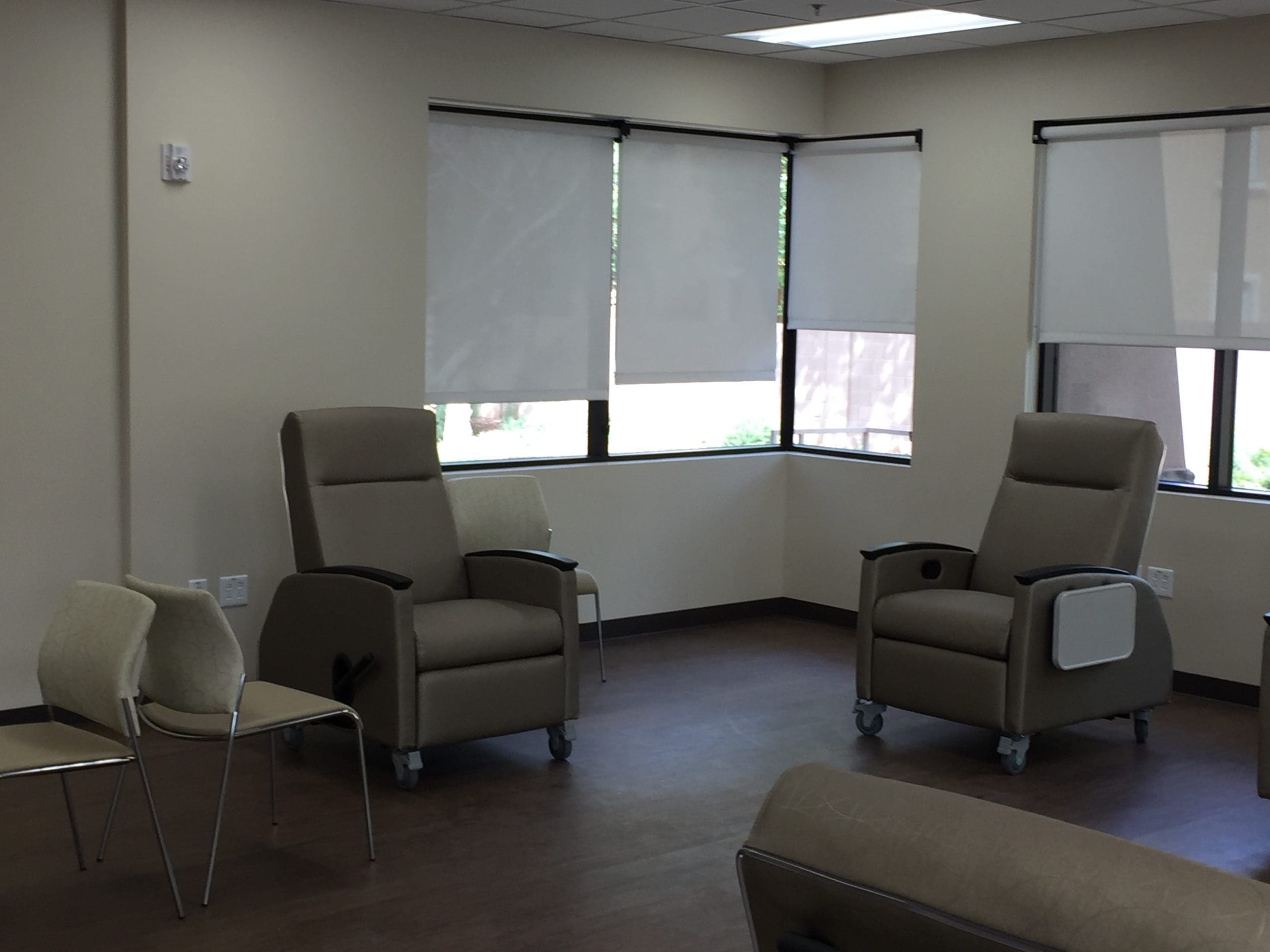 A room with beige reclining medical chairs on wheels, a couple of stackable chairs, large windows with blinds, and neutral-colored walls and flooring.