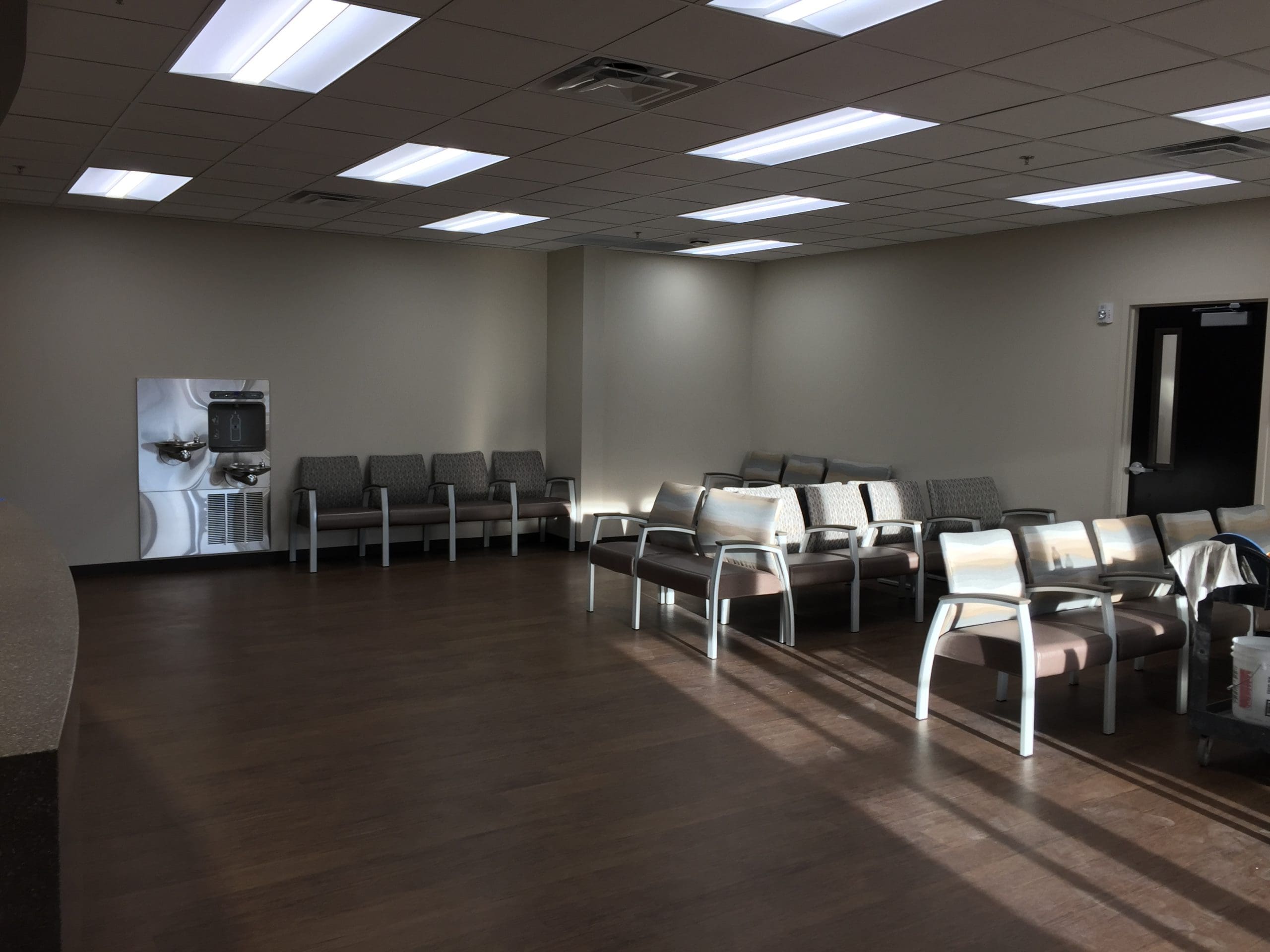 A waiting room with rows of empty chairs, a water fountain on the left wall, and wood flooring under bright overhead lighting.
